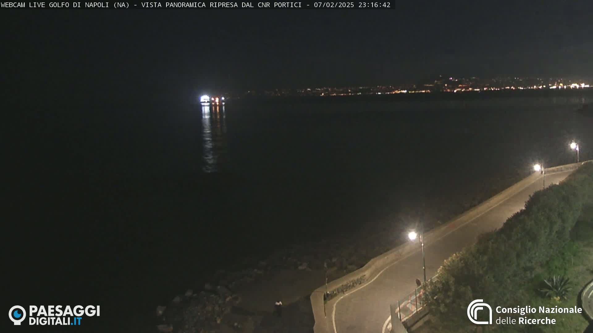 At night, a dark body of water reflects the lights of a distant city, a lone vessel is visible on the water, and a paved road curves alongside a low stone wall, lit by street lamps.