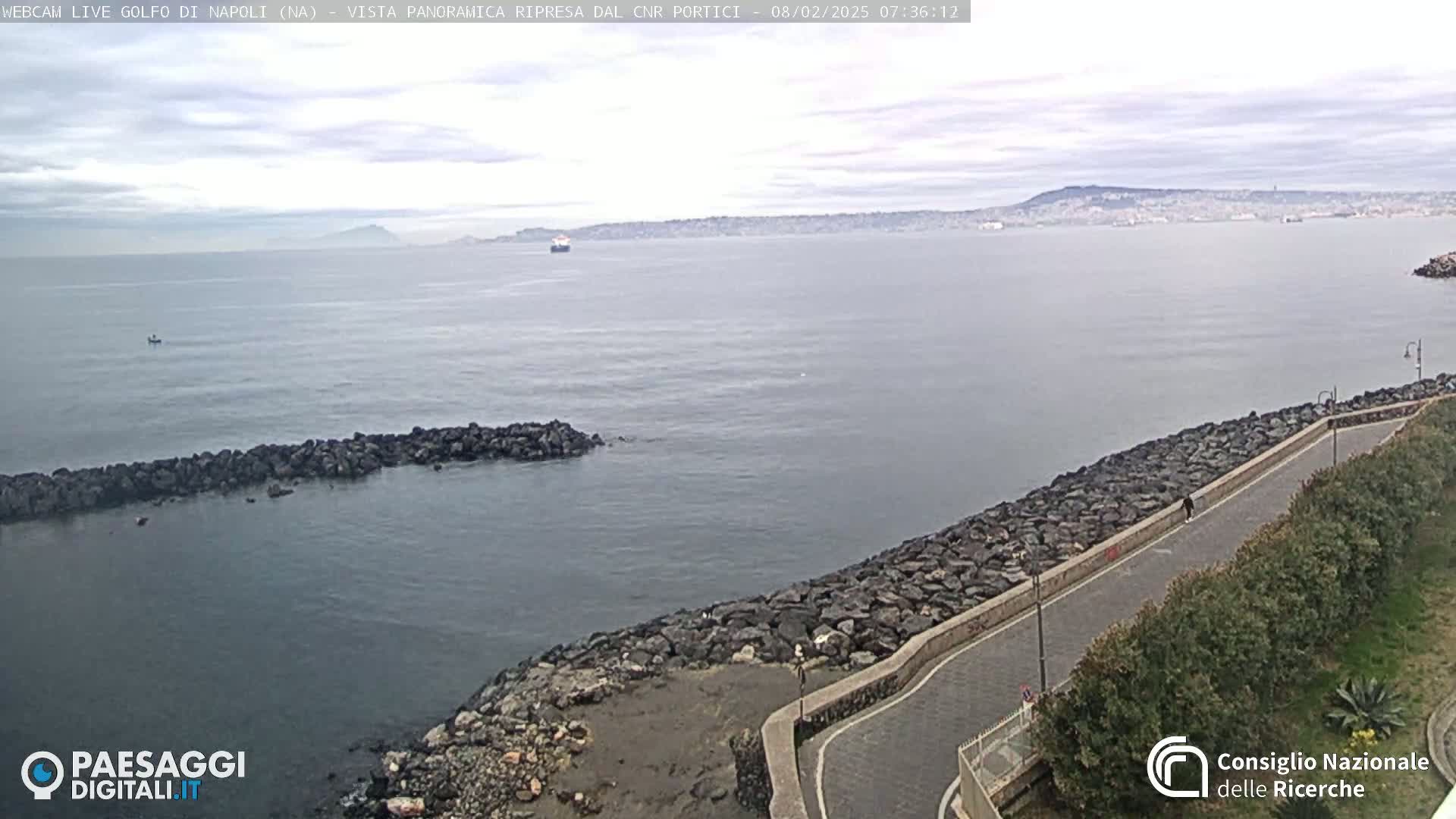A calm, overcast day shows a rocky shoreline with a paved path curving along the water's edge, overlooking a body of water with a distant mountain range and a lone vessel.