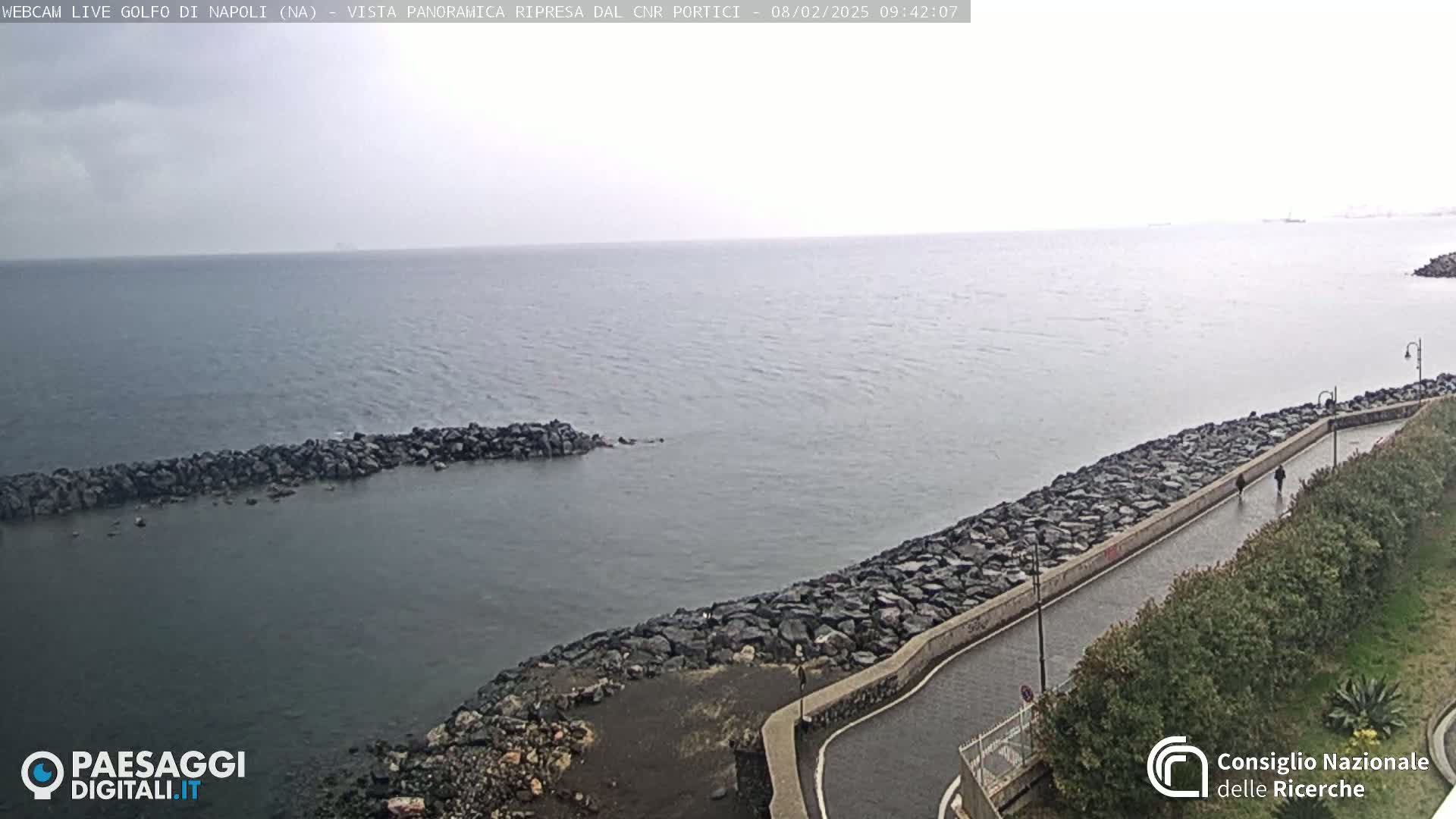 A paved walkway curves along a rocky shoreline overlooking a calm, gray sea under an overcast sky.