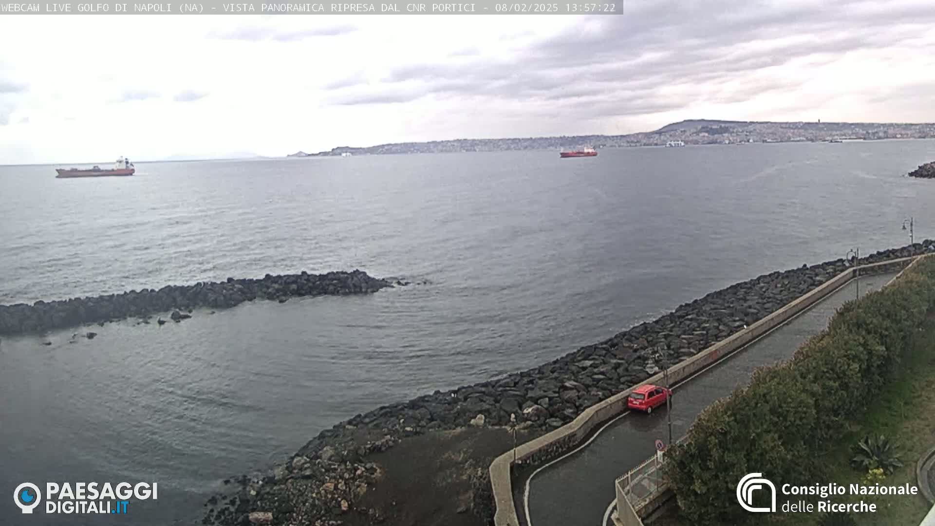 A partially cloudy day overlooking a calm body of water with a rocky shoreline, a red car, and several ships in the distance.