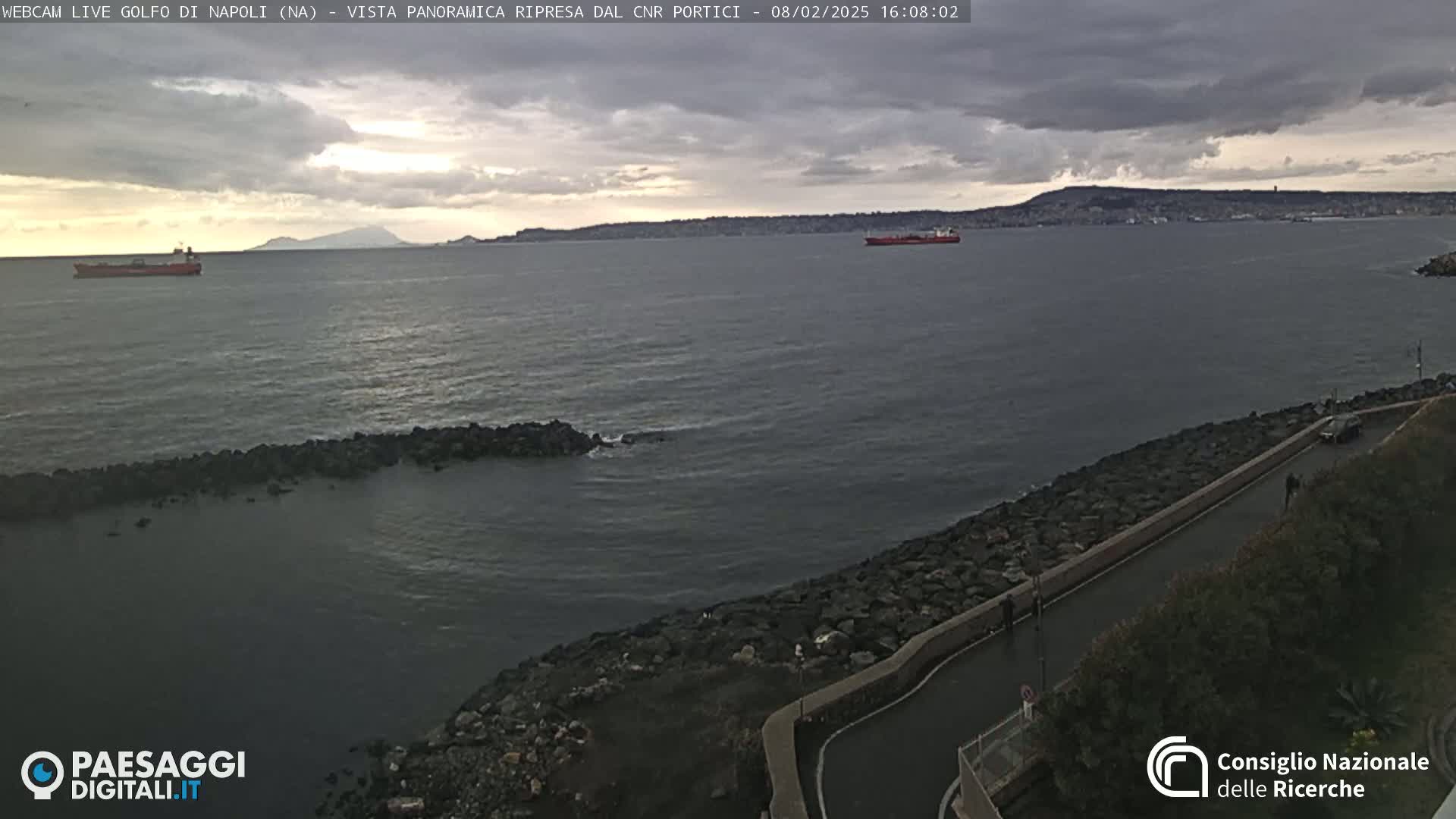 A partly cloudy day shows a rocky shoreline with a road next to a calm sea, with several ships visible in the distance.