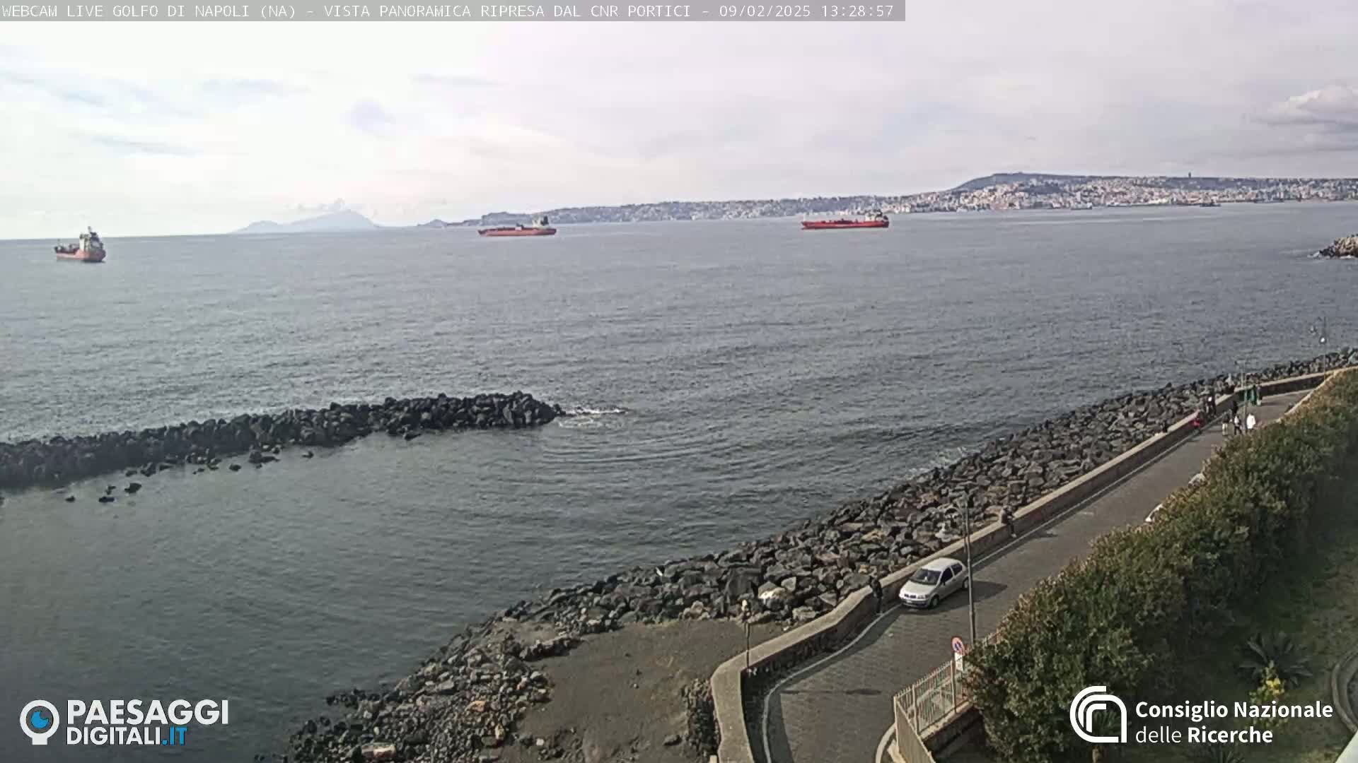 A partly cloudy day reveals a rocky coastline with a road and parked cars overlooking a calm sea with several ships and a distant mountain range.