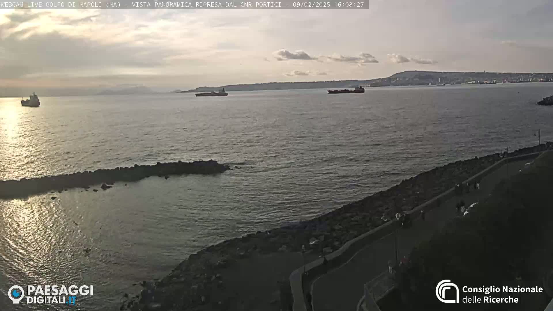 A mostly cloudy, dusky evening scene shows a calm body of water with several ships in the distance and a rocky shoreline with a paved walkway in the foreground.