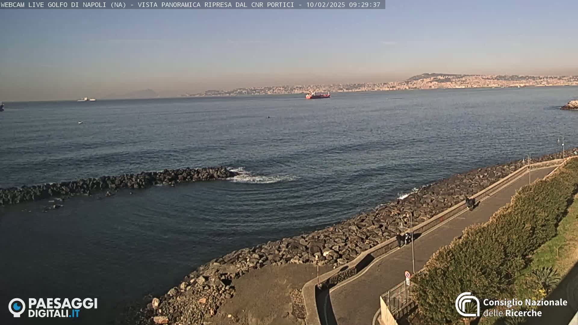 A rocky shoreline with a paved path curves gently beside a calm sea, under a clear sky, with a distant city and a large ship visible on the horizon.