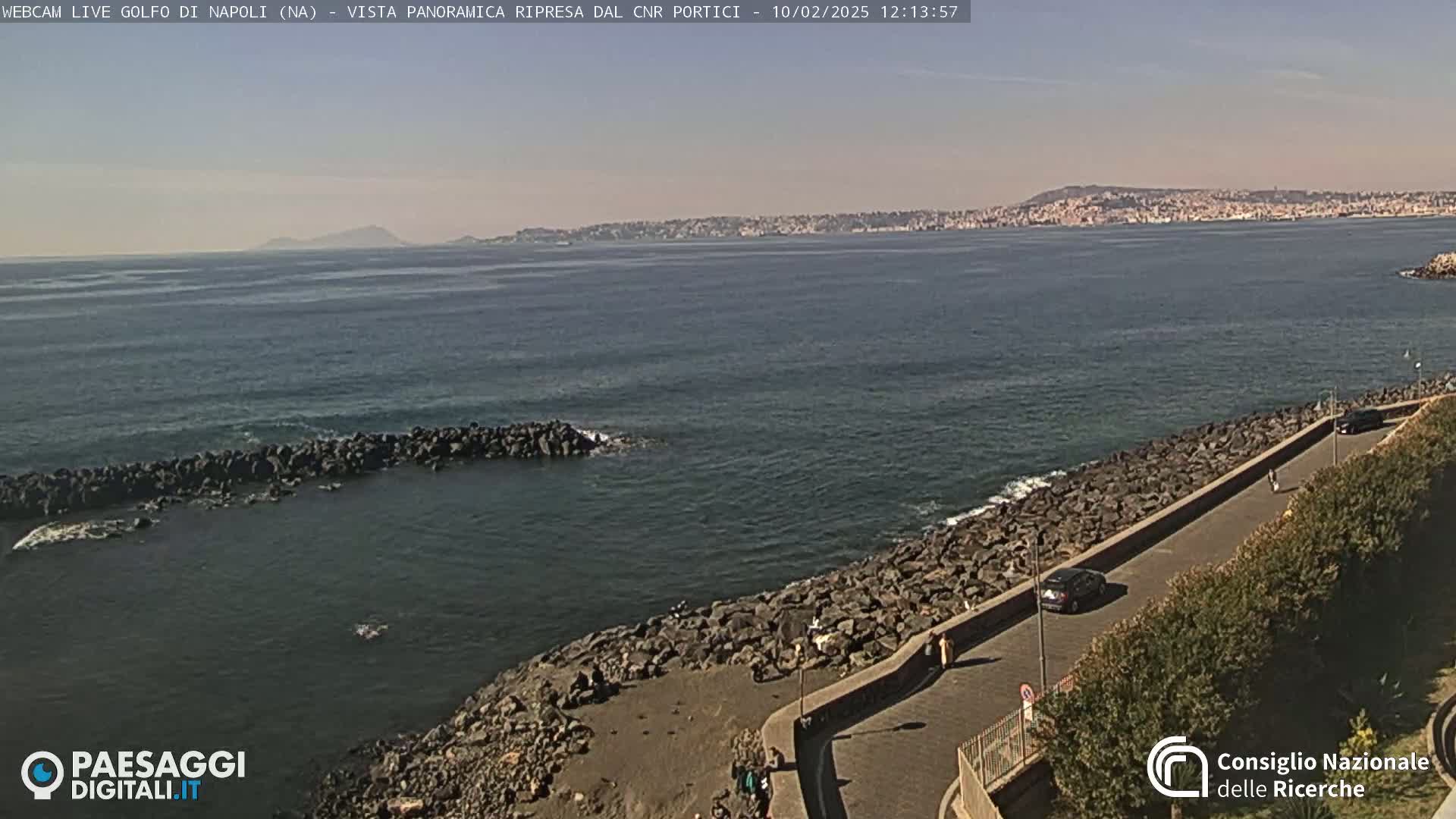 A panoramic view of a rocky coastline and calm sea under a clear sky shows a city in the distance.