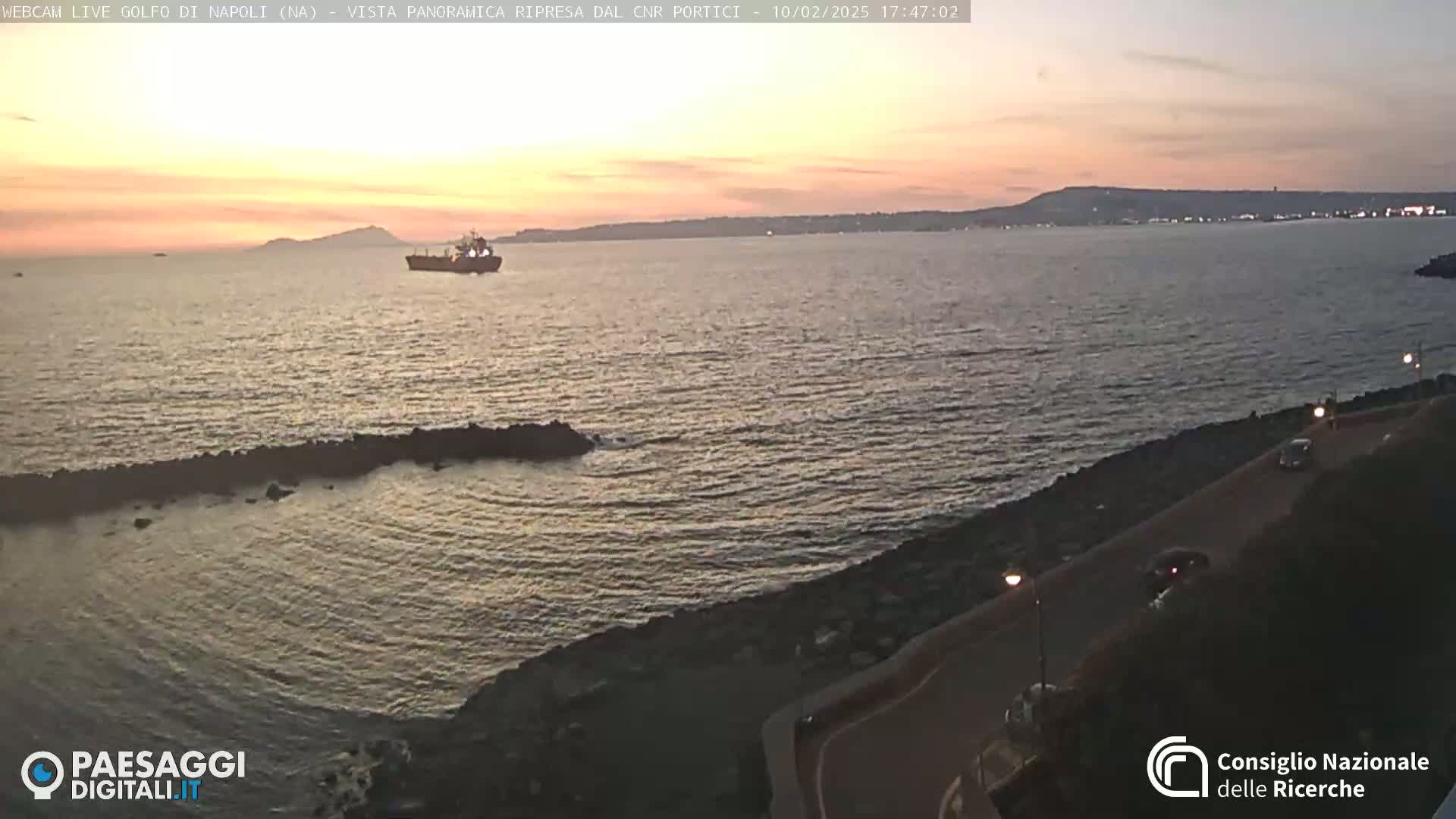 A large body of calm water at sunset is viewed from a coastal road, with a single ship visible in the distance.