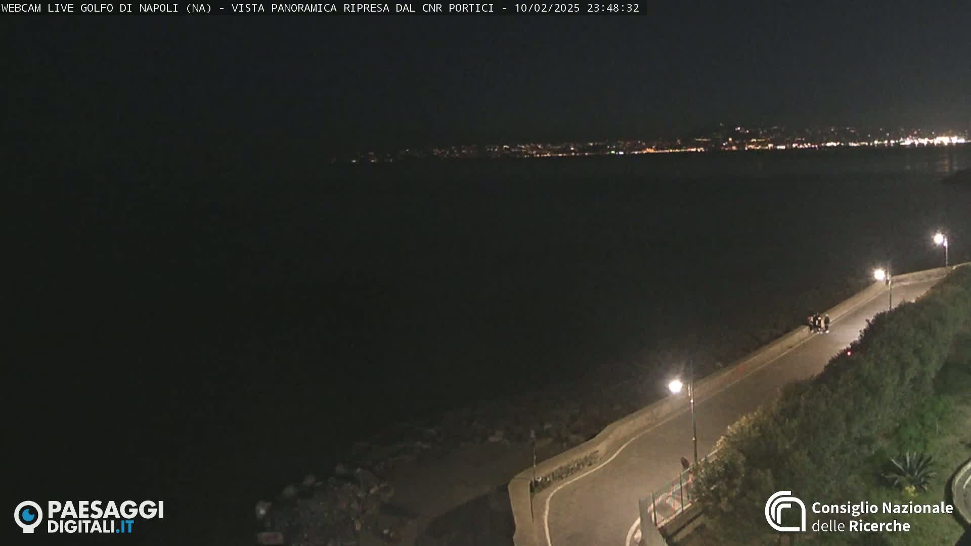 A dark nighttime view of a coastline with a city’s lights visible across the water, a road curving along the shoreline, and a few people standing near the edge.