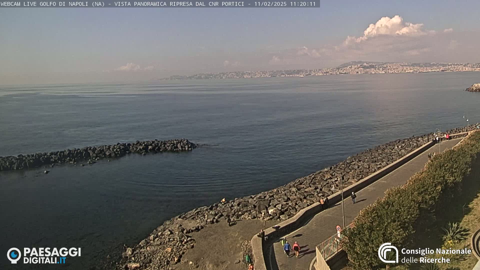A paved walkway lines a rocky shoreline overlooking a calm bay with a city visible in the distance under partly cloudy skies.