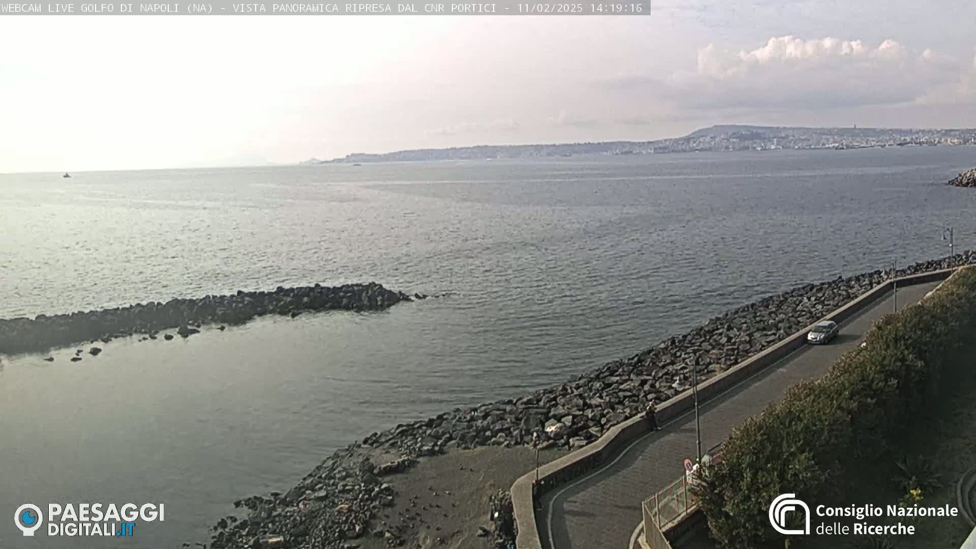 A rocky coastline curves gently alongside a calm body of water under a partly cloudy sky, with a distant city visible across the bay.