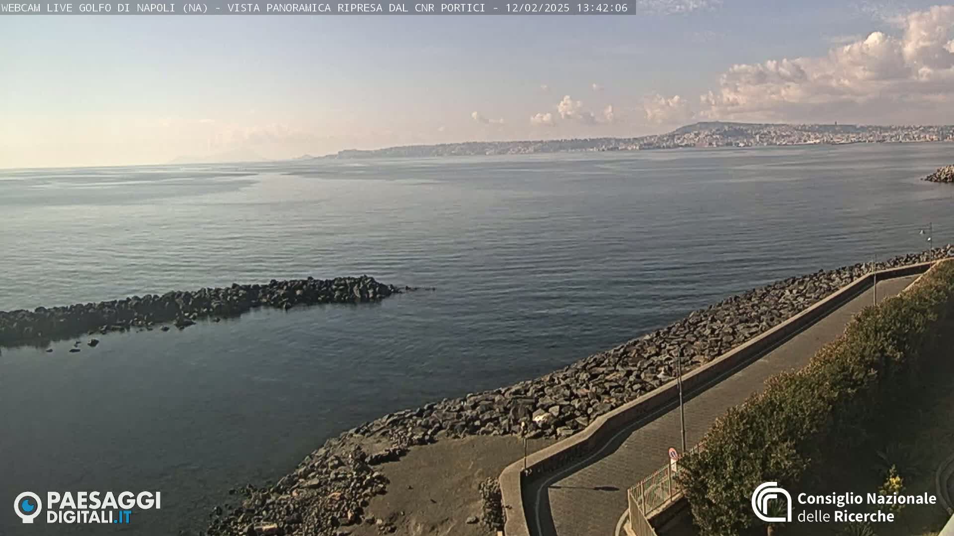 A partly cloudy day shows a rocky shoreline with a paved path beside a calm bay, a distant city skyline visible across the water.