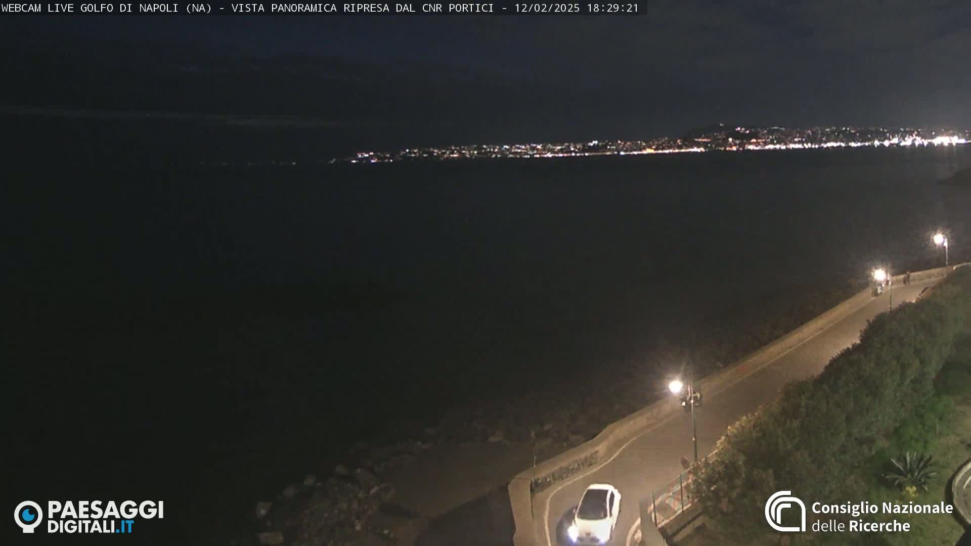 A nighttime, coastal scene shows a dark body of water with a distant, city skyline illuminated, and a roadway curving along the water's edge with a single car visible.
