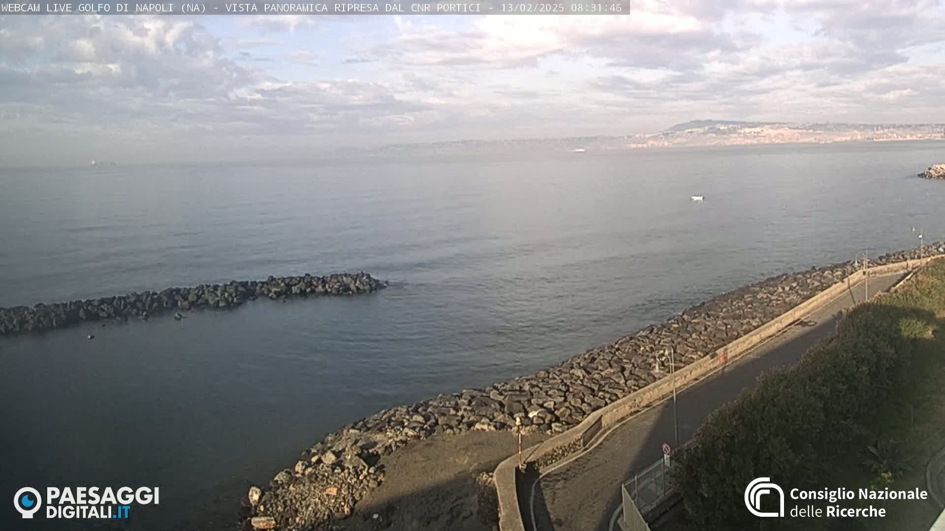 A calm sea with a rocky shoreline and a distant city under a partly cloudy sky.