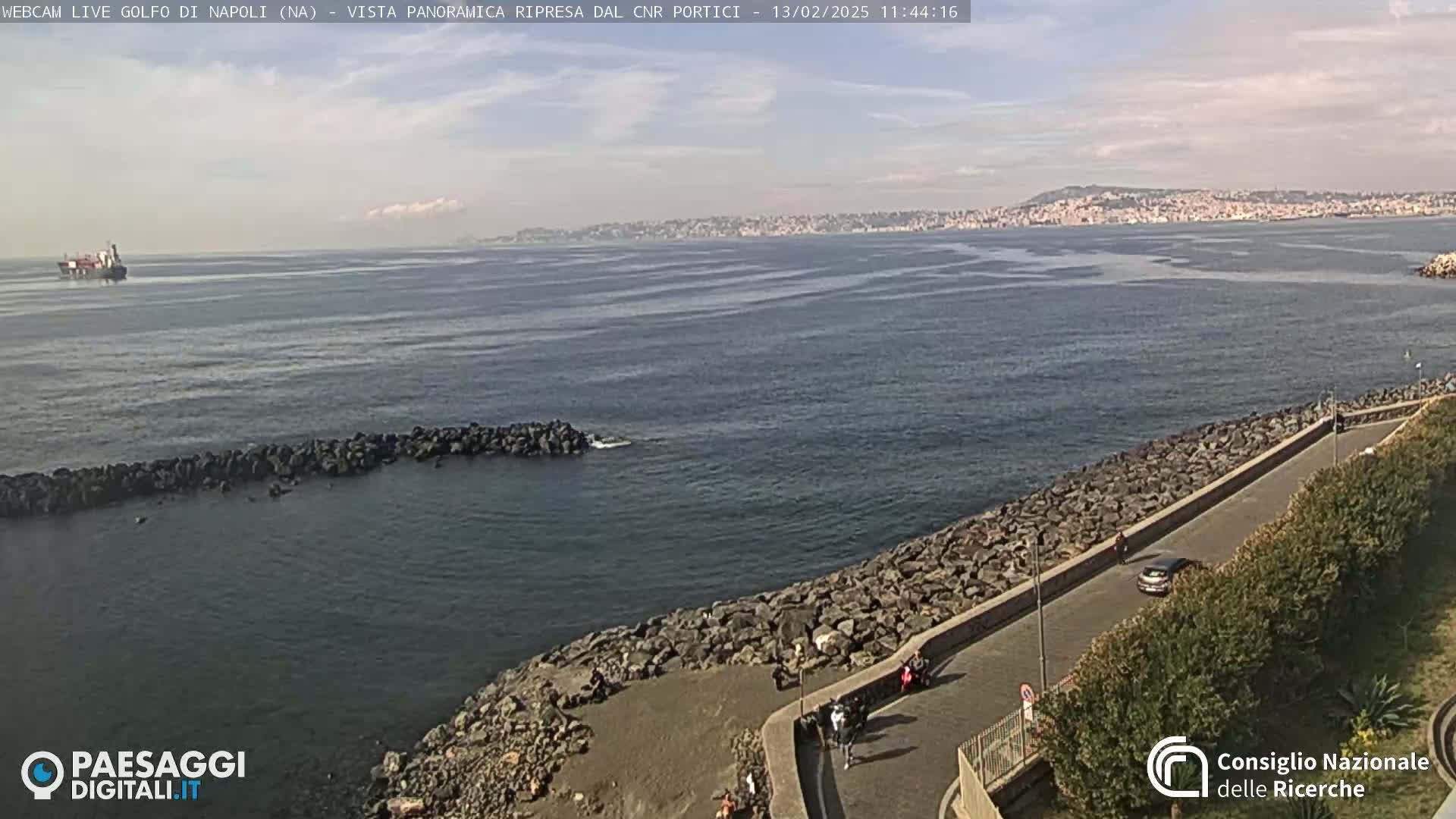 A rocky shoreline with a paved path and a few people walking, overlooking a calm body of water with a distant city skyline under a partly cloudy sky.