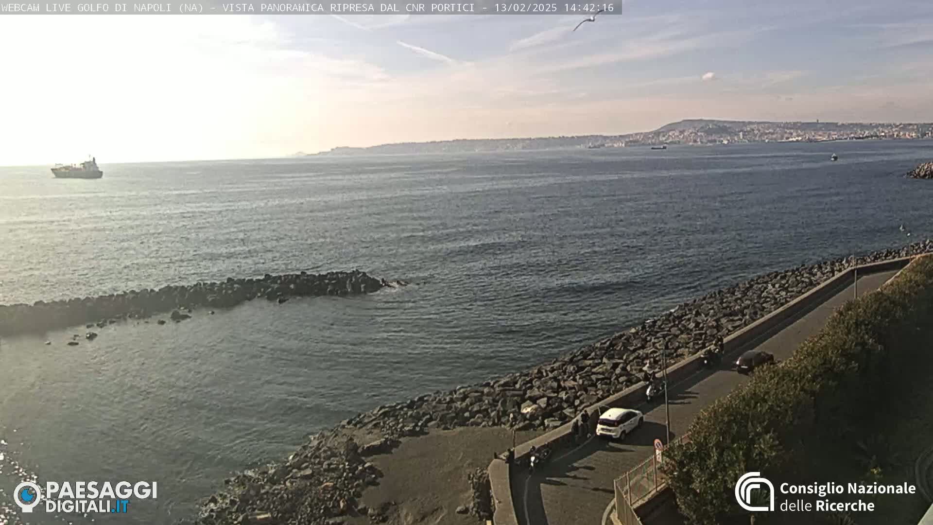 A mostly sunny, coastal view shows a calm sea with a distant city skyline, a rocky shoreline with a road and parked vehicles, and a single ship in the distance.