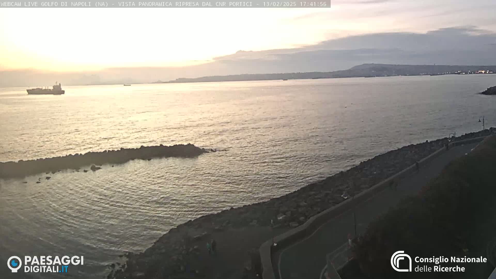 A calm sea at sunset is viewed from a rocky shoreline, with a distant landmass visible on the horizon.