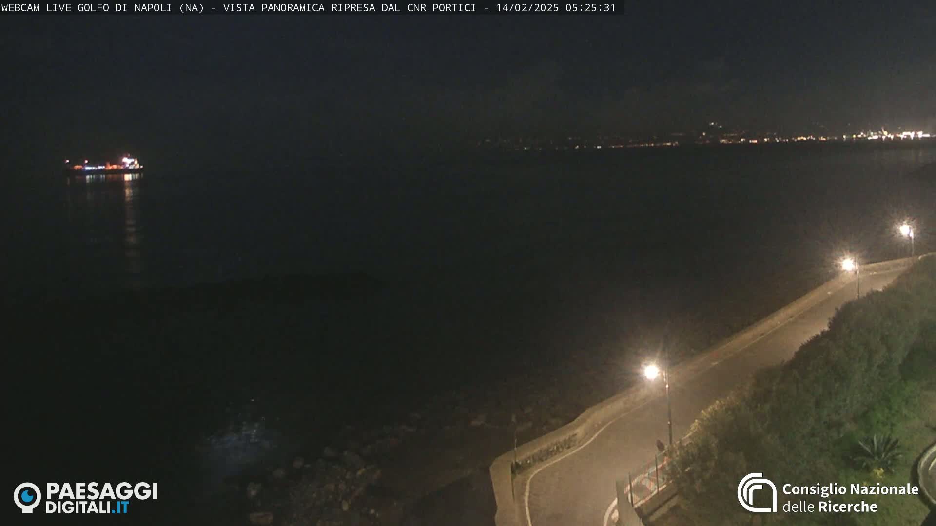 A nighttime, panoramic view of a dark body of water with a distant, lit city skyline and a single ship, alongside a road curving along the coast, lit by streetlamps.