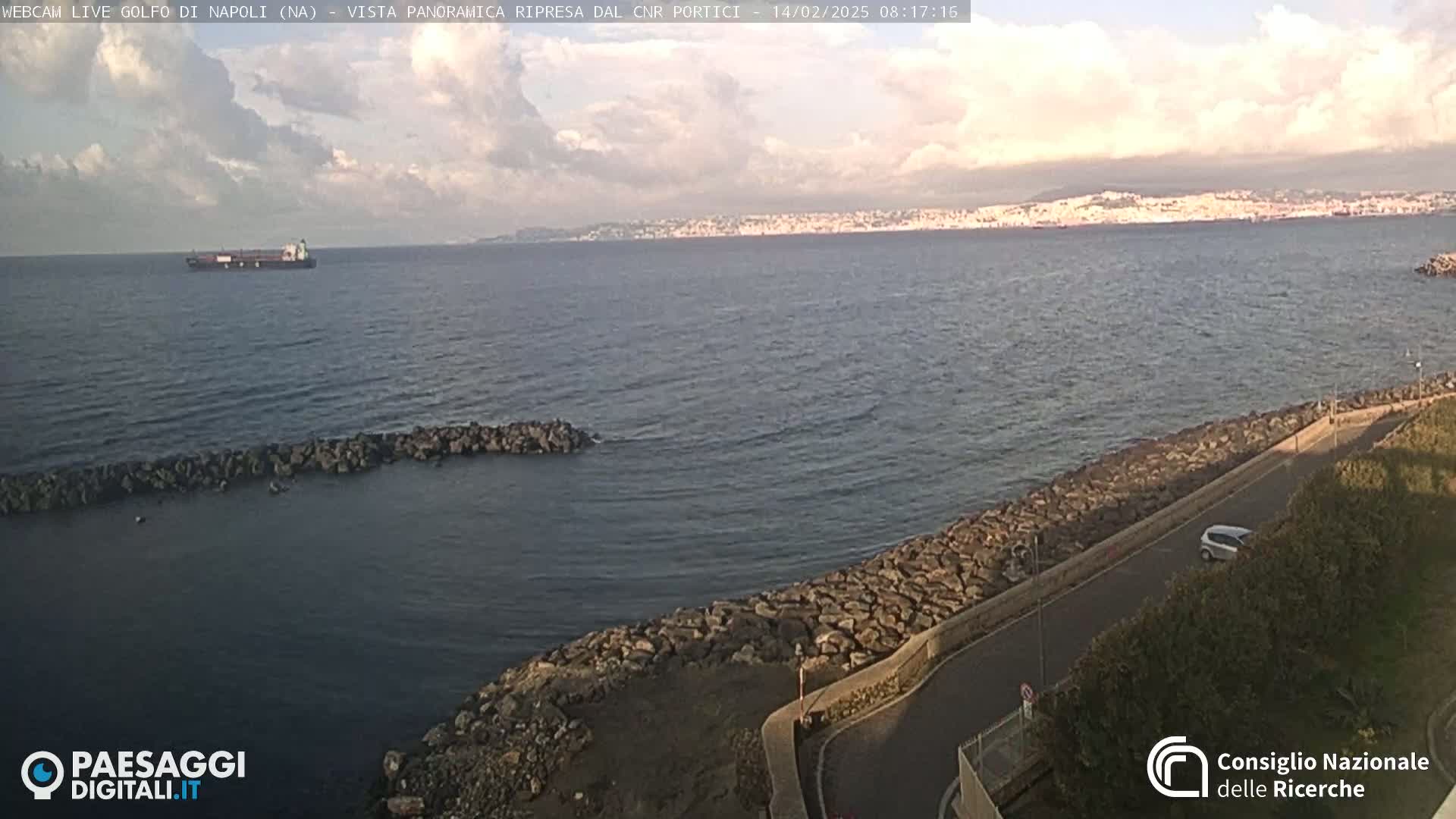 A partly cloudy day shows a large body of water with a distant cityscape, a cargo ship, and a rocky shoreline with a road next to it.