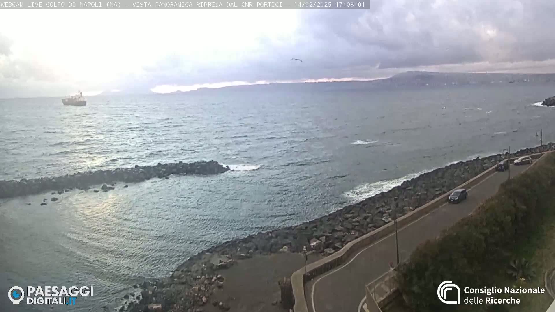 A partly cloudy day shows a rocky coastline with a road and several cars, overlooking a calm sea with a distant ship and mountains.