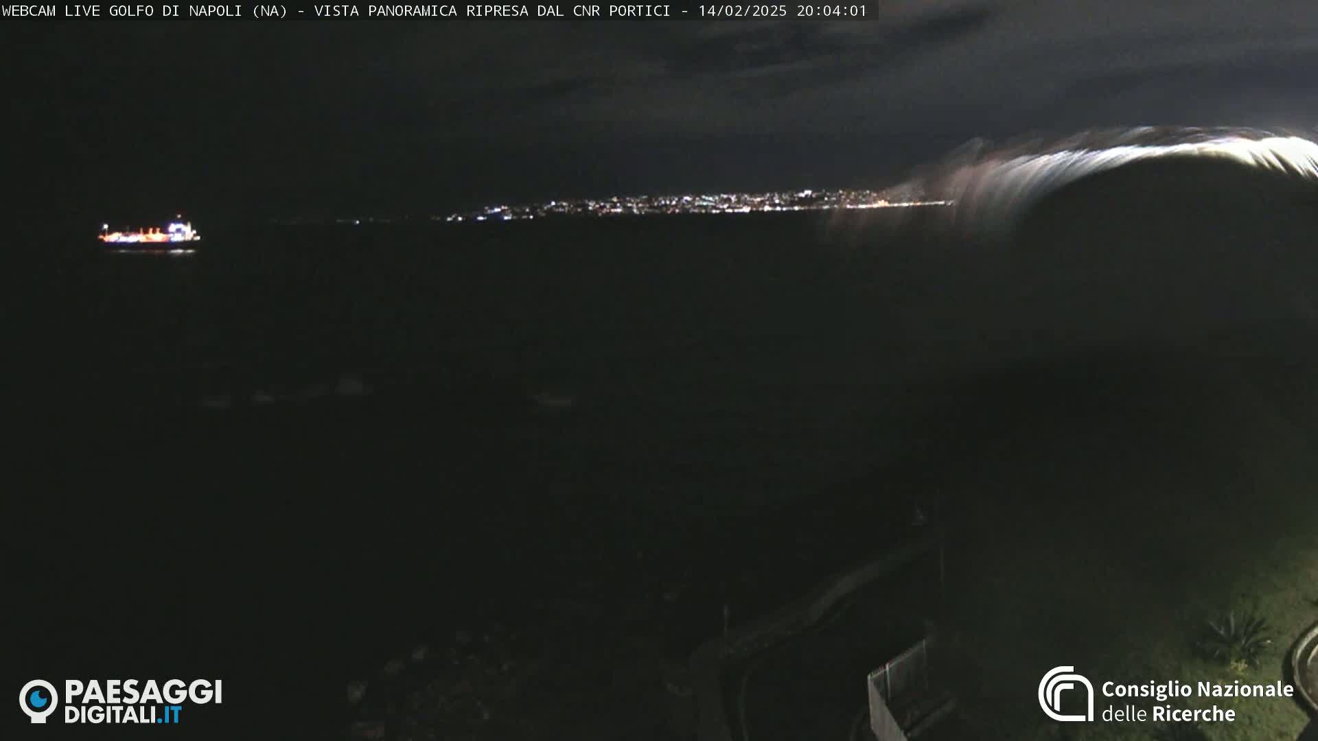 A nighttime, long shot shows a dark body of water with a distant city skyline illuminated by lights, a ship in the foreground, and a large wave breaking in the right corner.