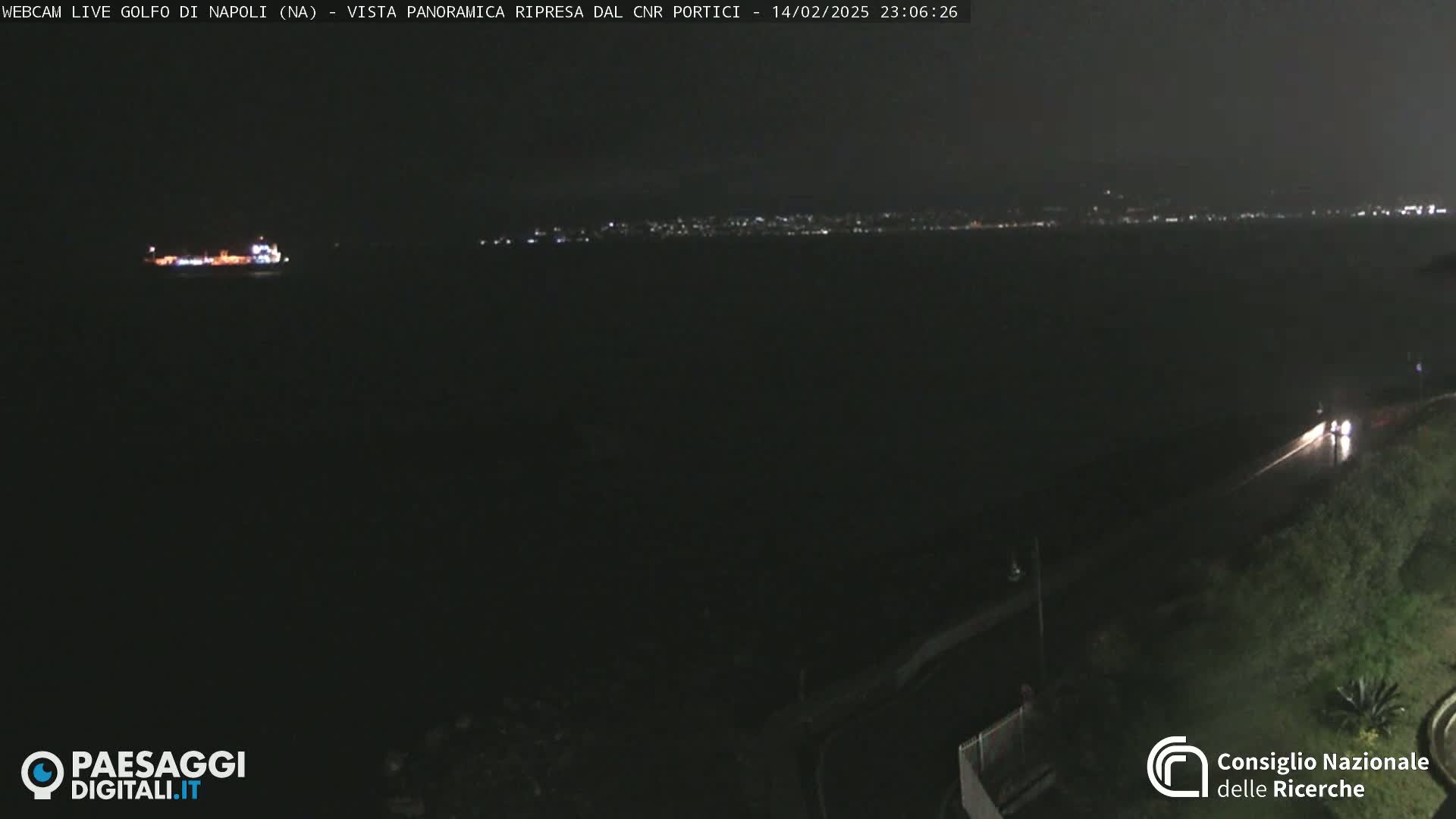 A nighttime, long shot shows a dark body of water with a distant city skyline faintly lit, a large ship in the water, and a road with a few vehicles visible in the foreground.