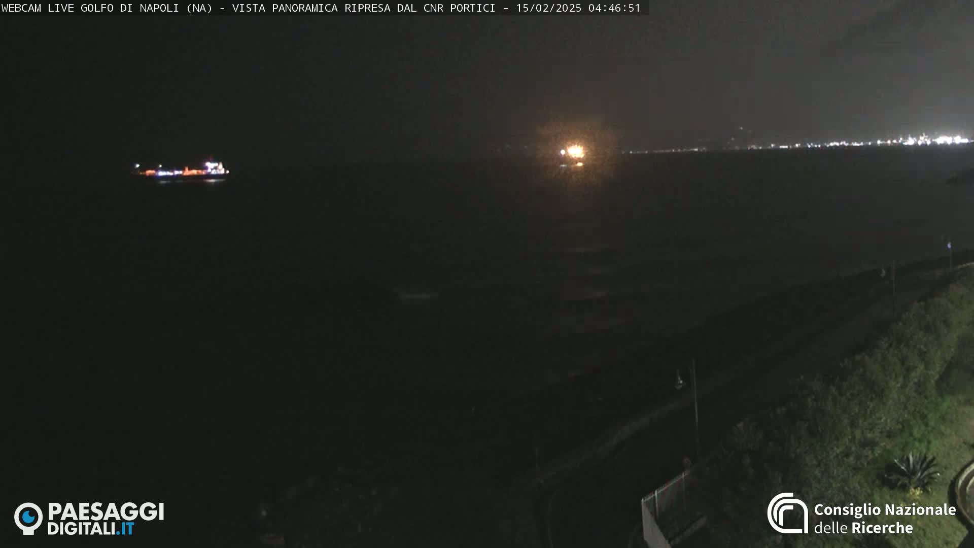 A nighttime, panoramic view of a dark body of water with a distant shoreline showing lights and a single ship.