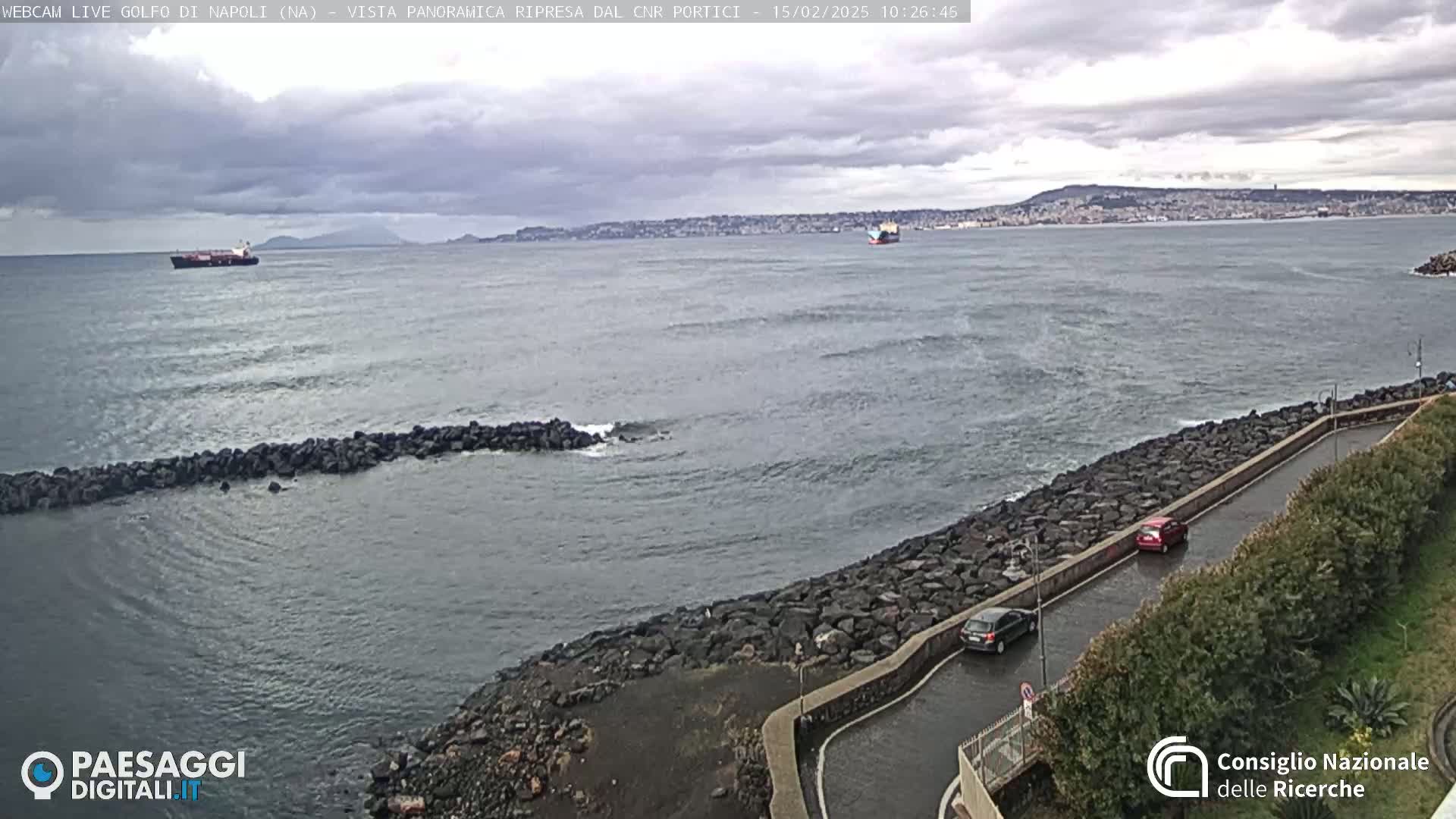 A rocky shoreline with a road and some cars overlooks a choppy sea with a few ships visible in the distance under a cloudy sky.