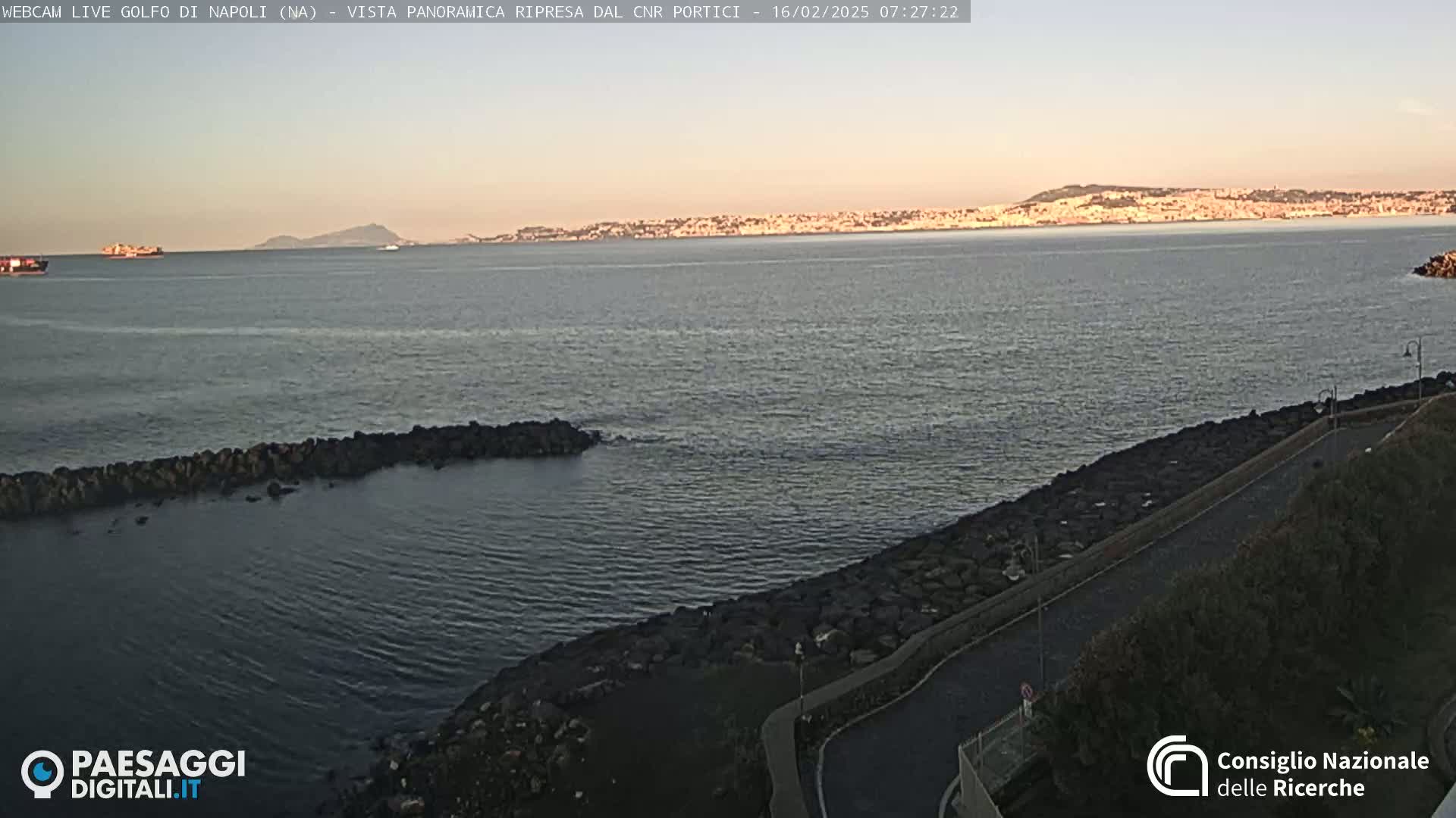 A calm sea with a rocky shoreline and a distant city skyline under a clear, dusky sky.