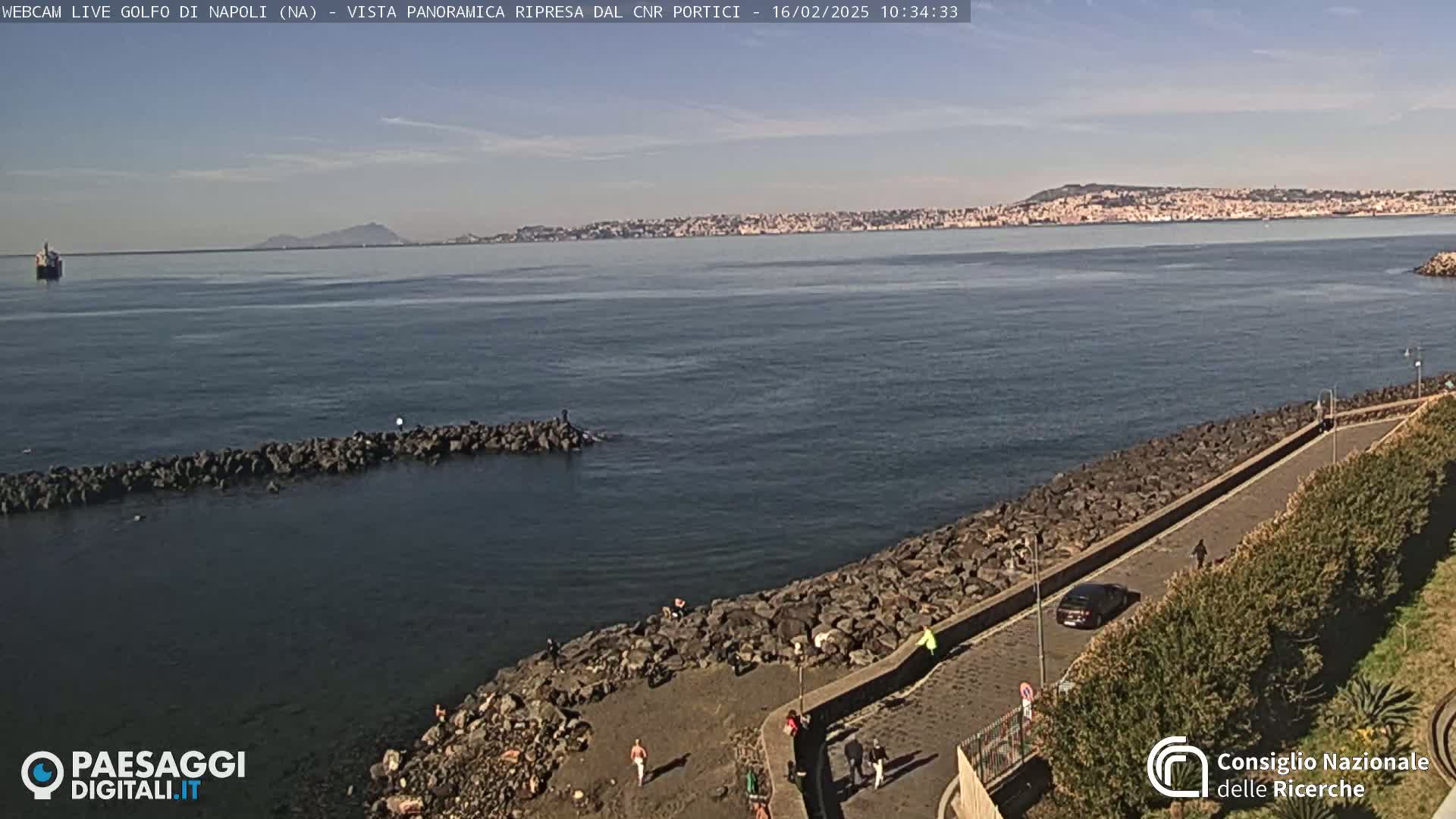 A rocky shoreline with people walking along a paved path overlooks a calm bay, with a distant city and mountains visible under a clear sky.