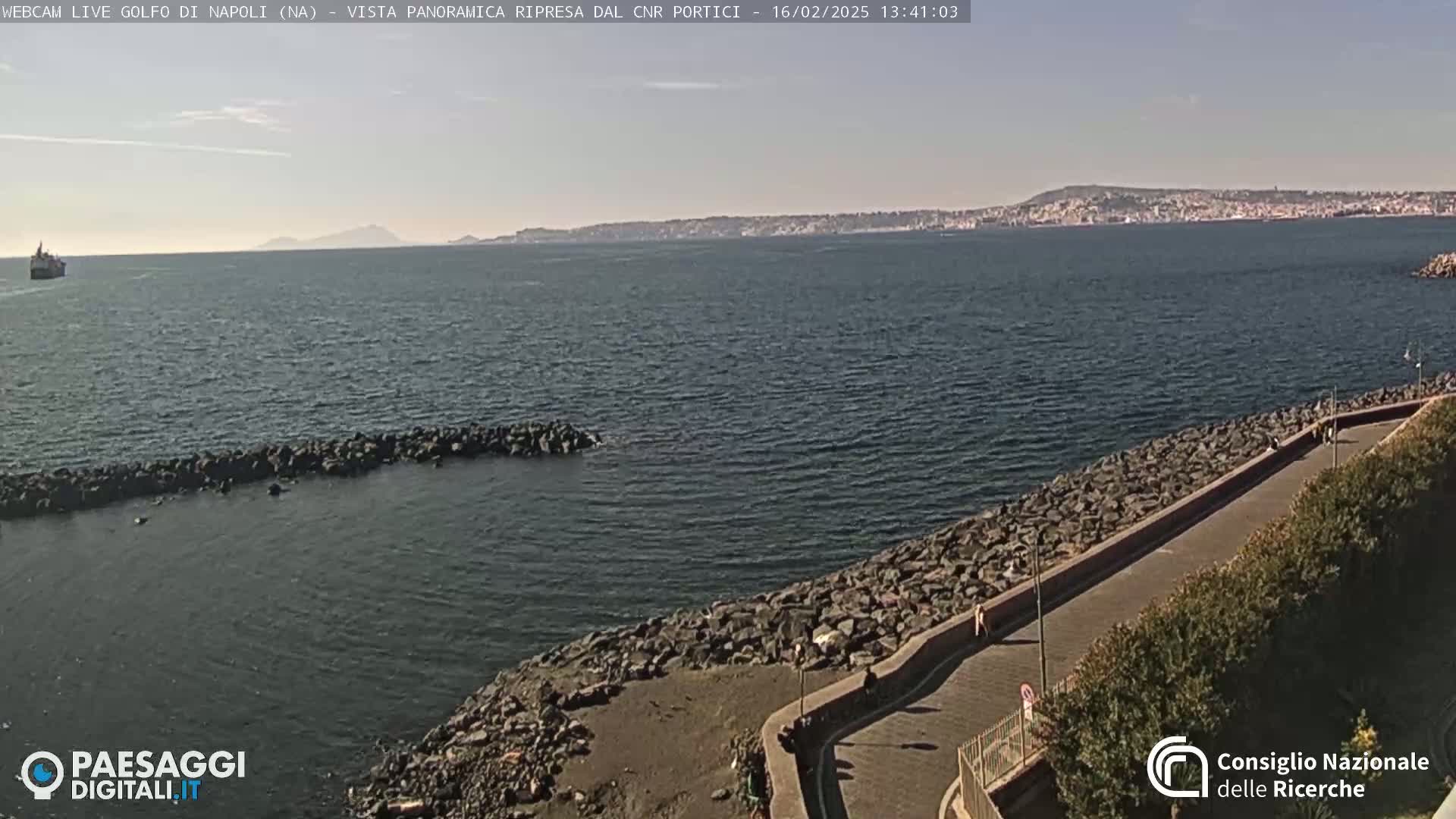 A rocky shoreline with a paved walkway overlooks a calm bay, under a mostly clear sky, with a distant cityscape visible across the water.