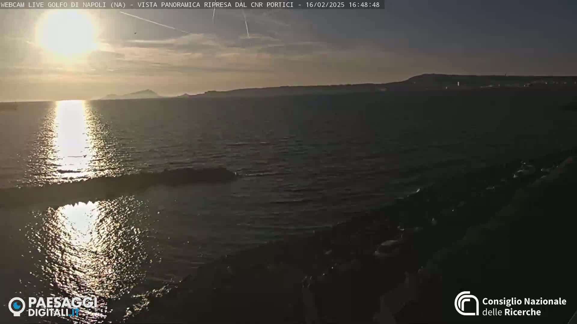 A calm sea reflects the setting sun, with a distant shoreline visible under a mostly clear sky.