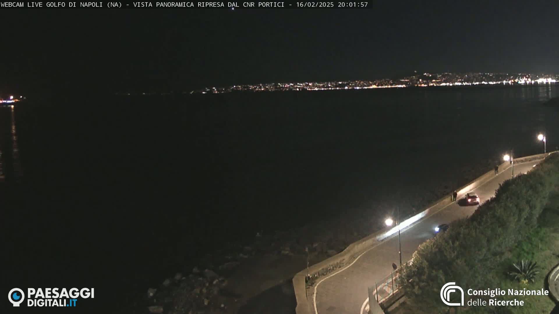A nighttime view of a dark body of water with a distant city skyline illuminated by lights, and a paved road curving along the water's edge with a few cars and people visible.