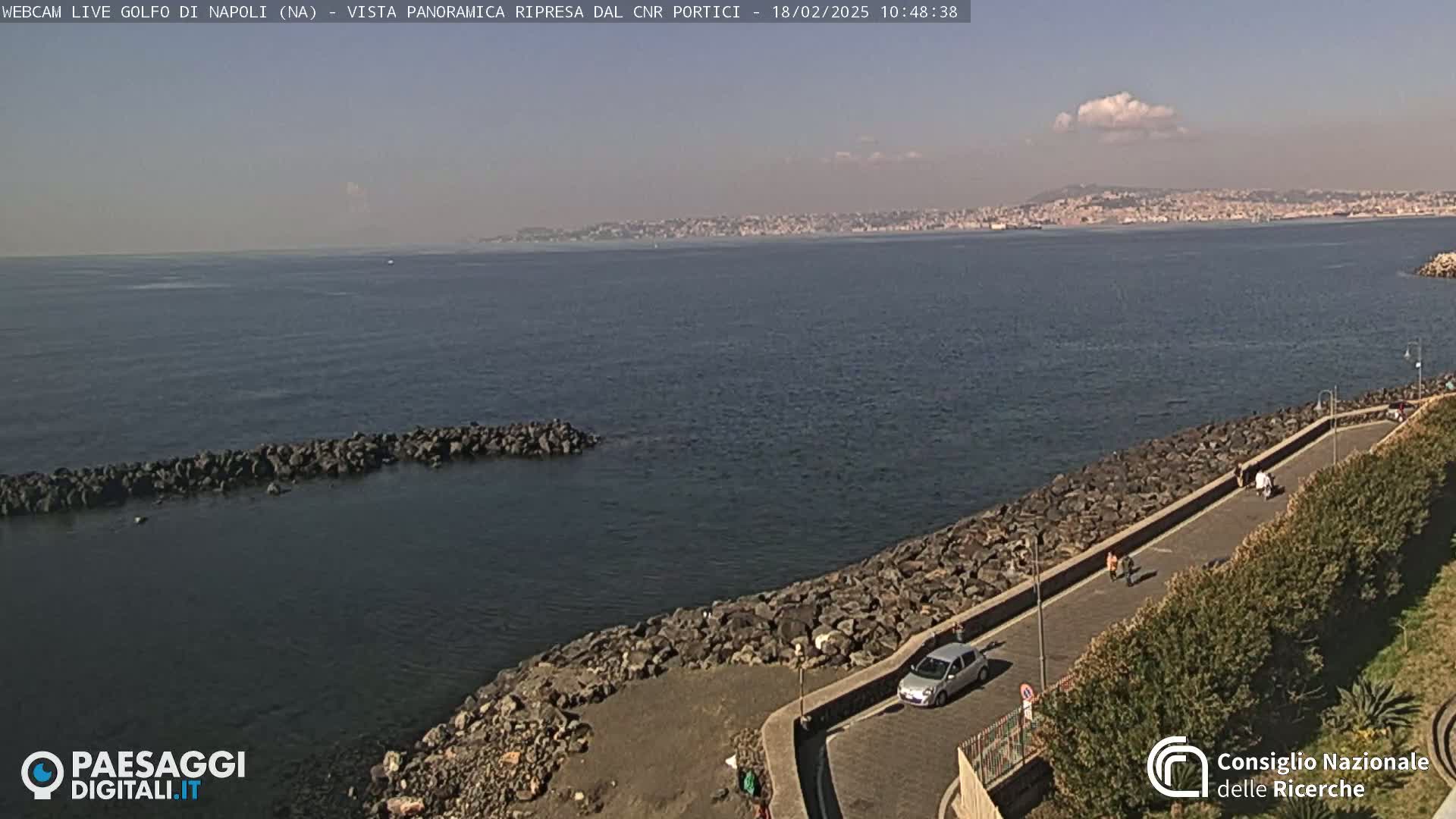 A partly cloudy day shows a rocky coastline with a paved walkway, a few people strolling, and a car parked; in the distance, a city is visible across a calm body of water.