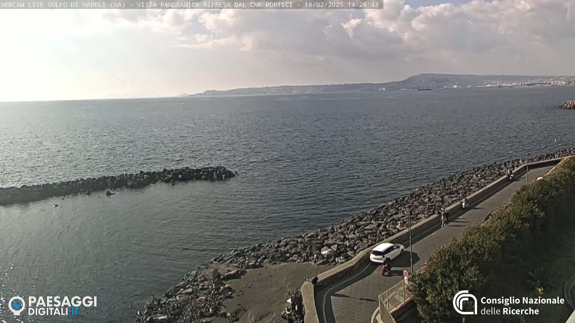 A partly cloudy day reveals a panoramic view of a calm sea with a rocky shoreline, a road with several vehicles and people, and distant land in the background.