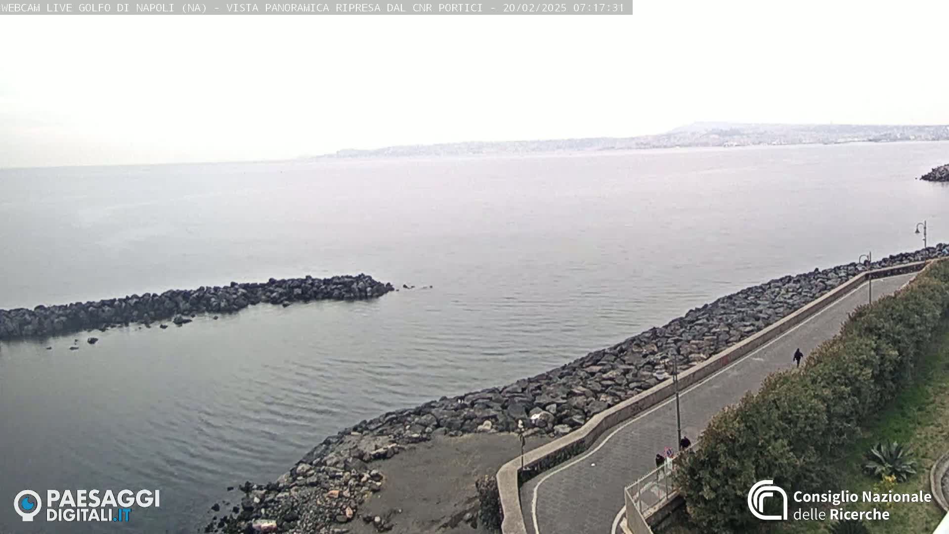 A rocky shoreline with a paved path curves alongside calm gray water under an overcast sky, extending to a distant hazy coastline.