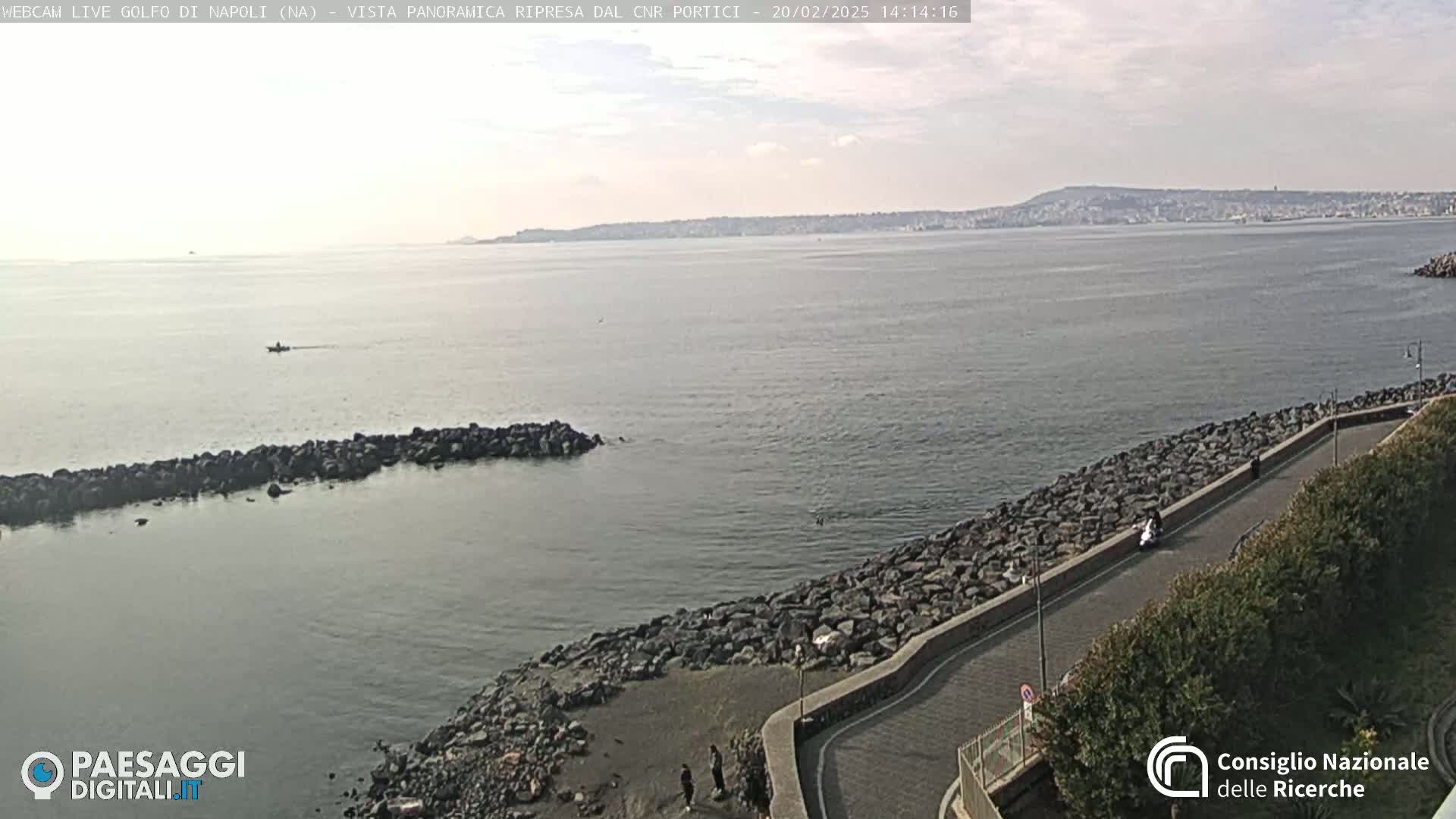 A rocky shoreline curves gently around a calm bay, under a mostly clear sky, with a distant city visible across the water.