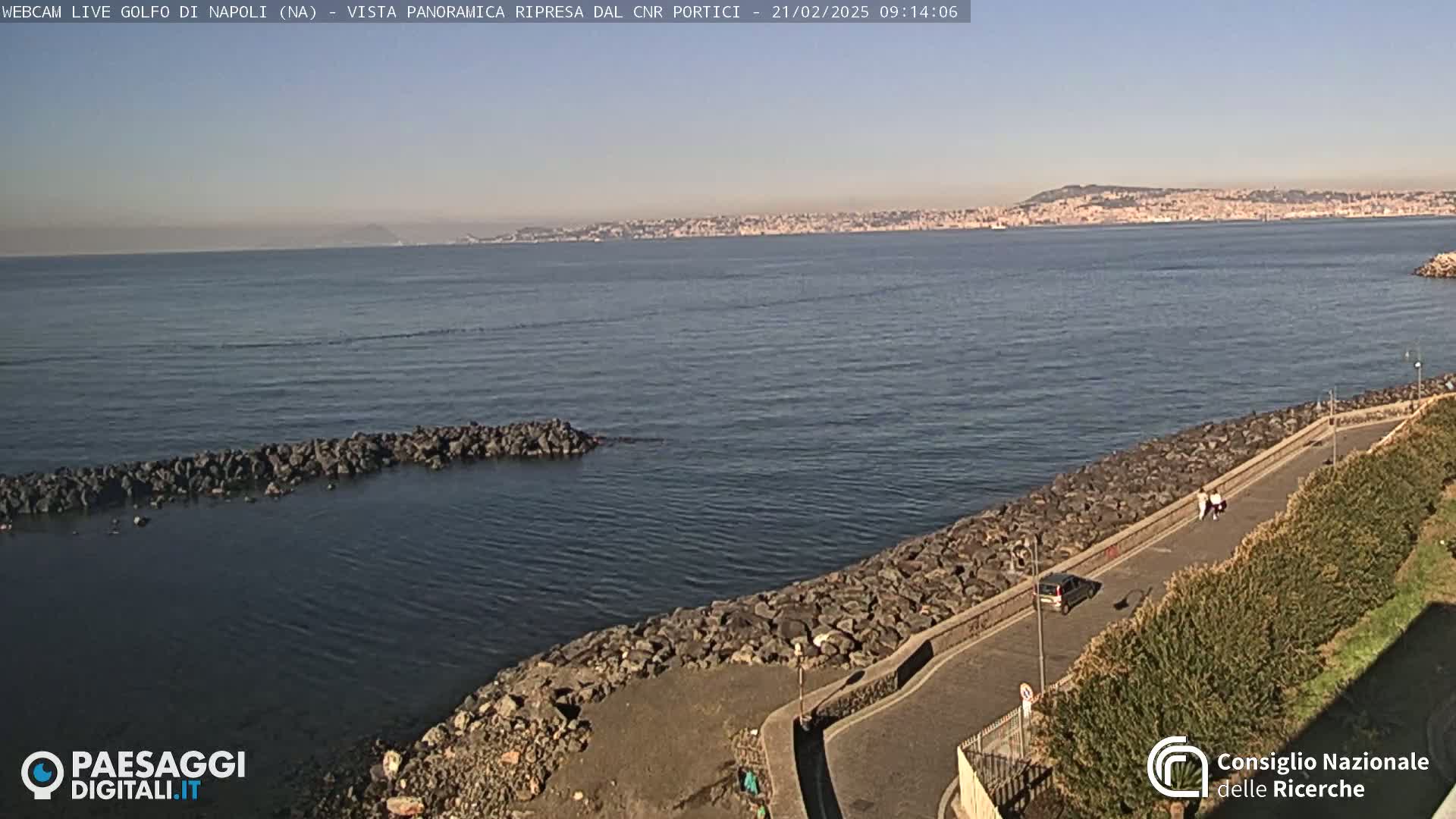 A rocky shoreline with a paved path curves alongside calm, dark-blue water, offering a view of a distant city skyline under a clear, sunny sky.