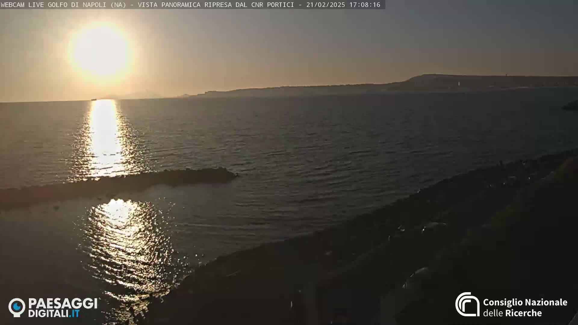 A setting sun reflects on calm water near a rocky shoreline, with a distant landmass visible on the horizon.
