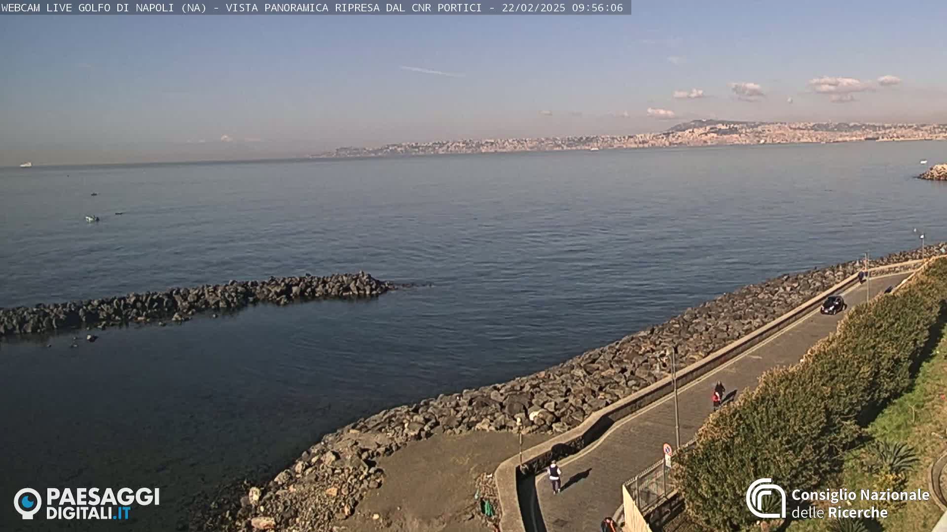A rocky shoreline curves along a calm bay, under a mostly clear sky, with a distant city visible across the water.