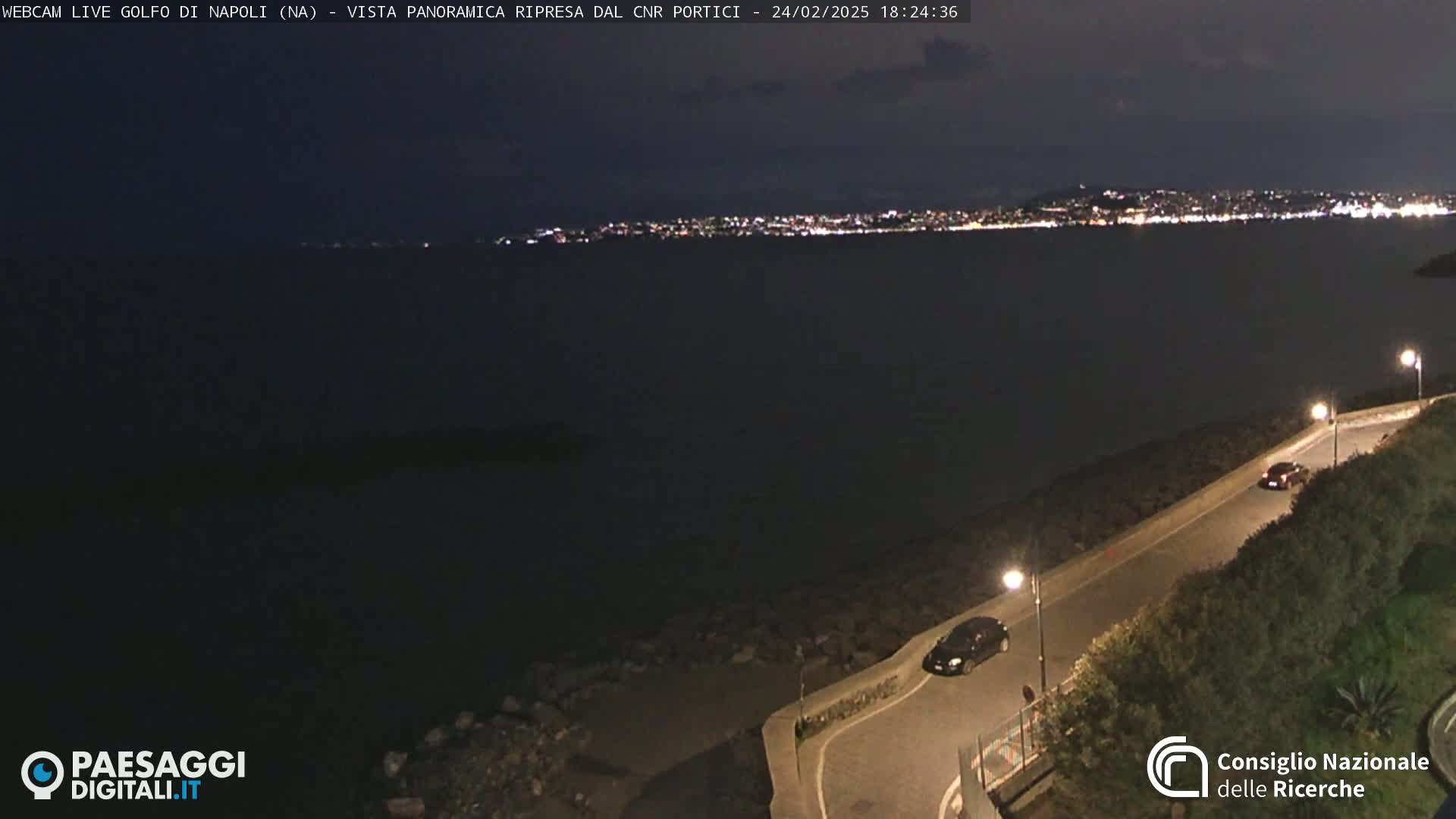 A nighttime, panoramic view shows a dark body of water with a city's lights in the distance, a road curving along a shoreline, and a few cars visible.