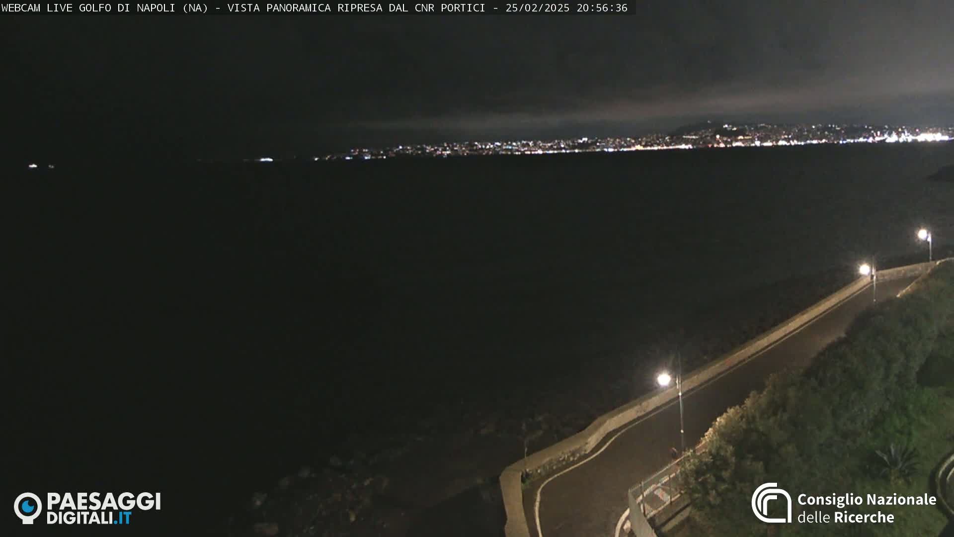 A nighttime, panoramic view shows a dark body of water with a brightly lit city in the distance, and a dark roadway curving along a waterfront in the foreground.