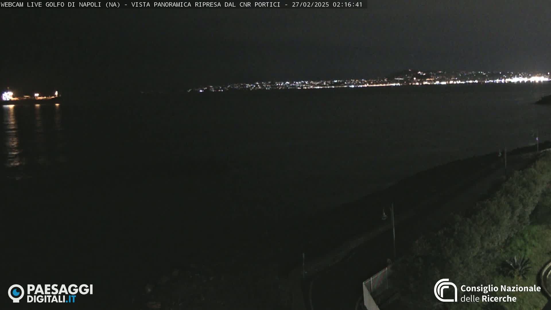 A nighttime, panoramic view of a dark body of water with a distant city skyline illuminated by lights.