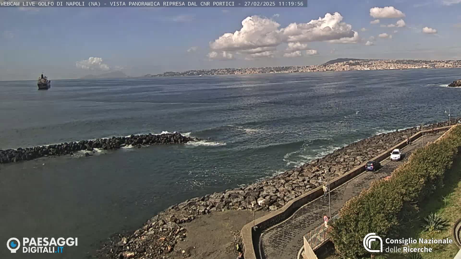 A partly cloudy day shows a cargo ship at sea, a rocky shoreline with a road and parked cars, and a city in the distance.