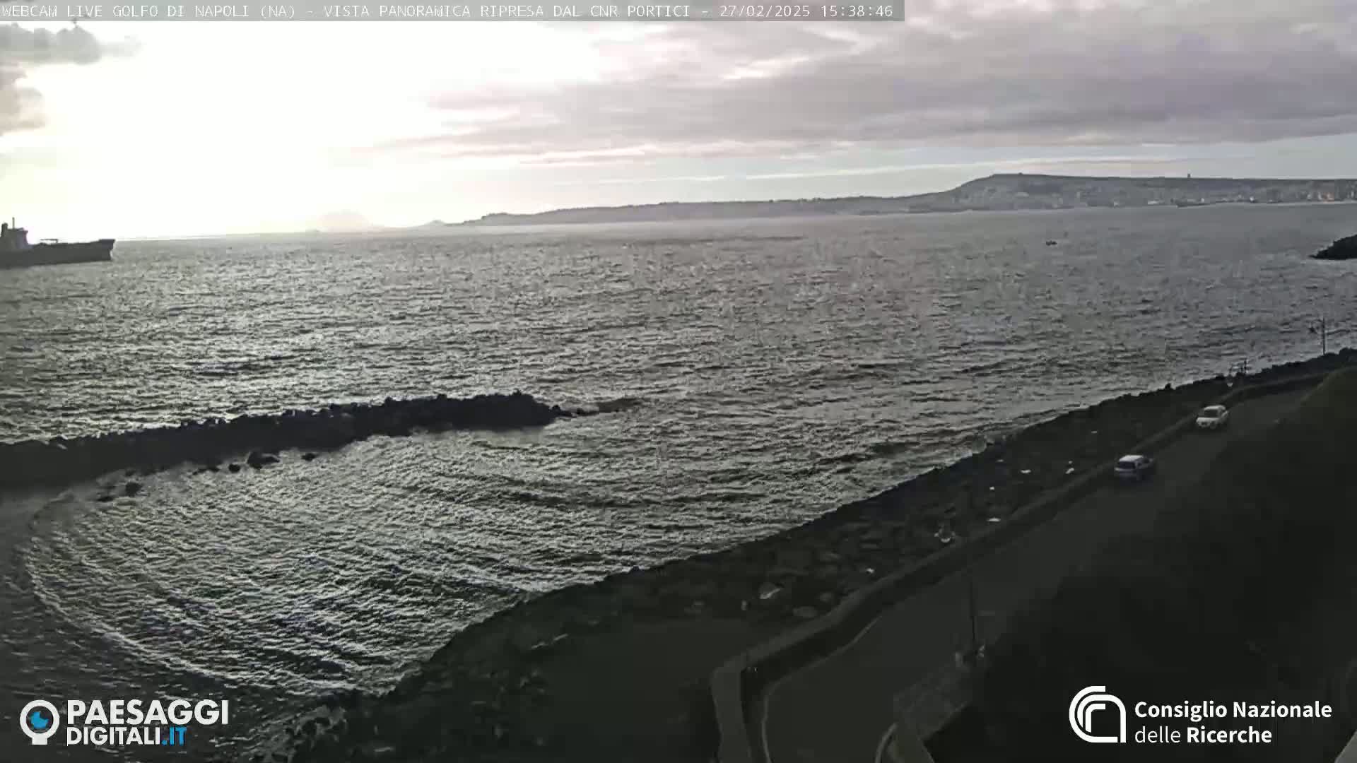 A partly cloudy day shows a view of a calm sea with a distant coastline and a few cars driving on a road next to the water.