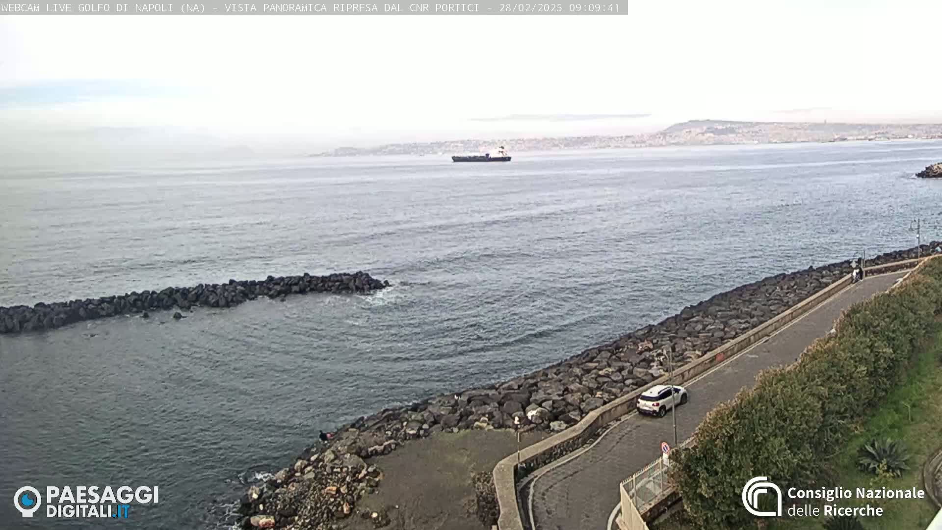 A hazy, overcast day reveals a coastal road curving along a rocky shoreline, overlooking a calm sea with a distant cargo ship and a hazy mountain range in the background.