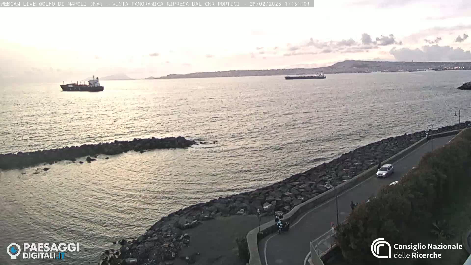 A calm, twilight sea with two cargo ships in the distance, viewed from a rocky shoreline with a road curving along the coast.