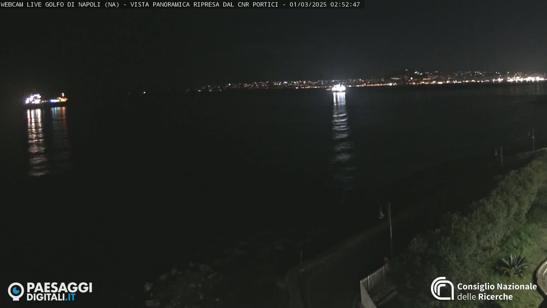 At night, a dark body of water reflects the lights of a distant city and two illuminated vessels.