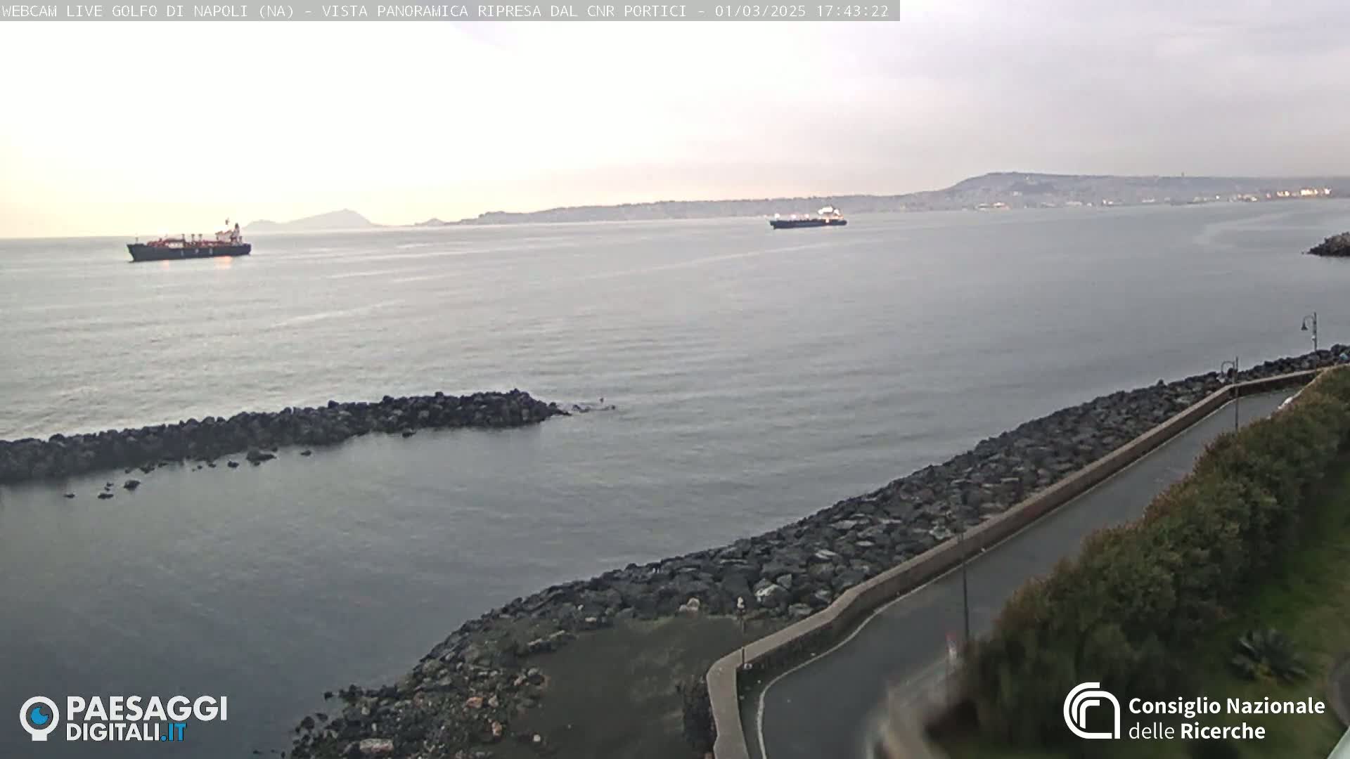 A calm, overcast day shows two ships in a bay, with a rocky shoreline and a road visible in the foreground.