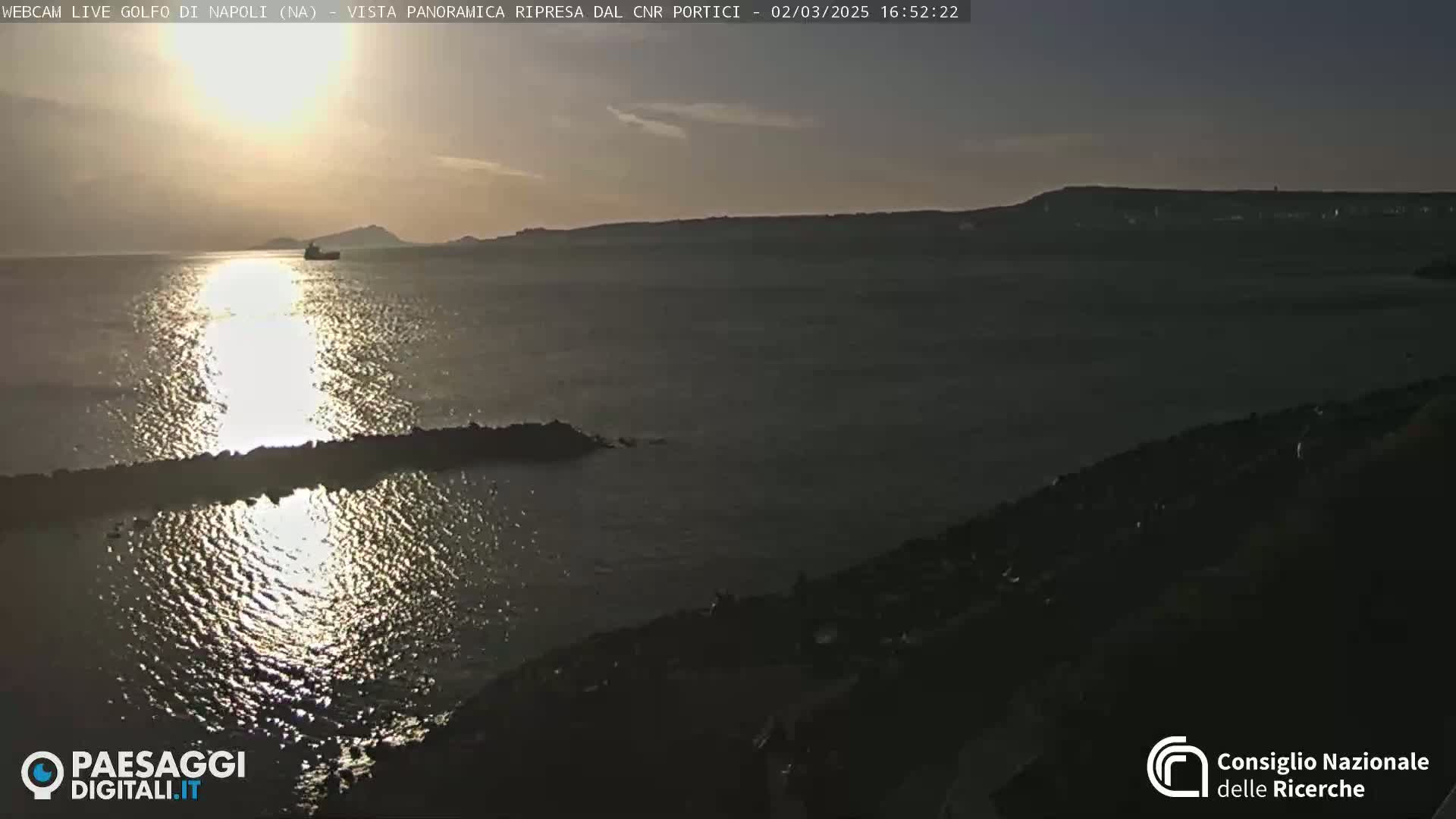 A calm body of water reflects the setting sun, with a distant shoreline and a single vessel visible on the horizon.