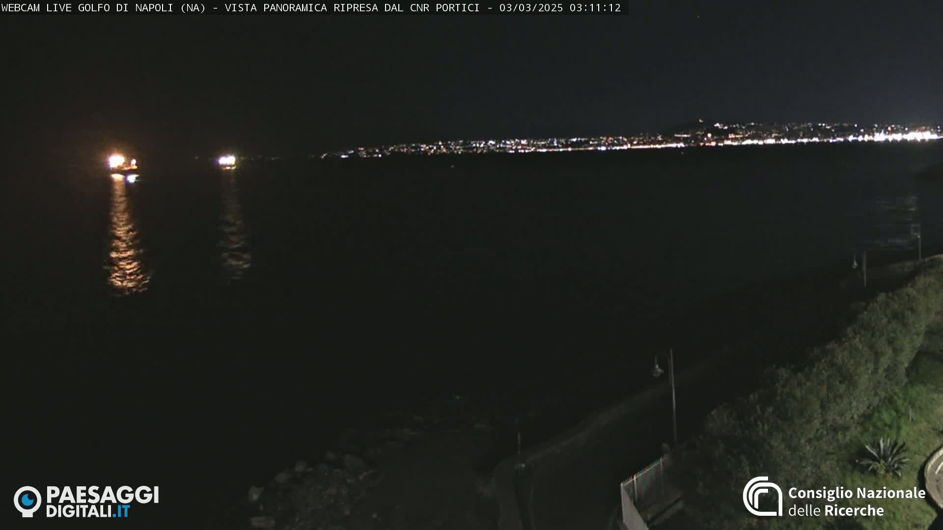 At night, a dark body of water reflects the lights of a distant city skyline, with two illuminated ships visible in the foreground.