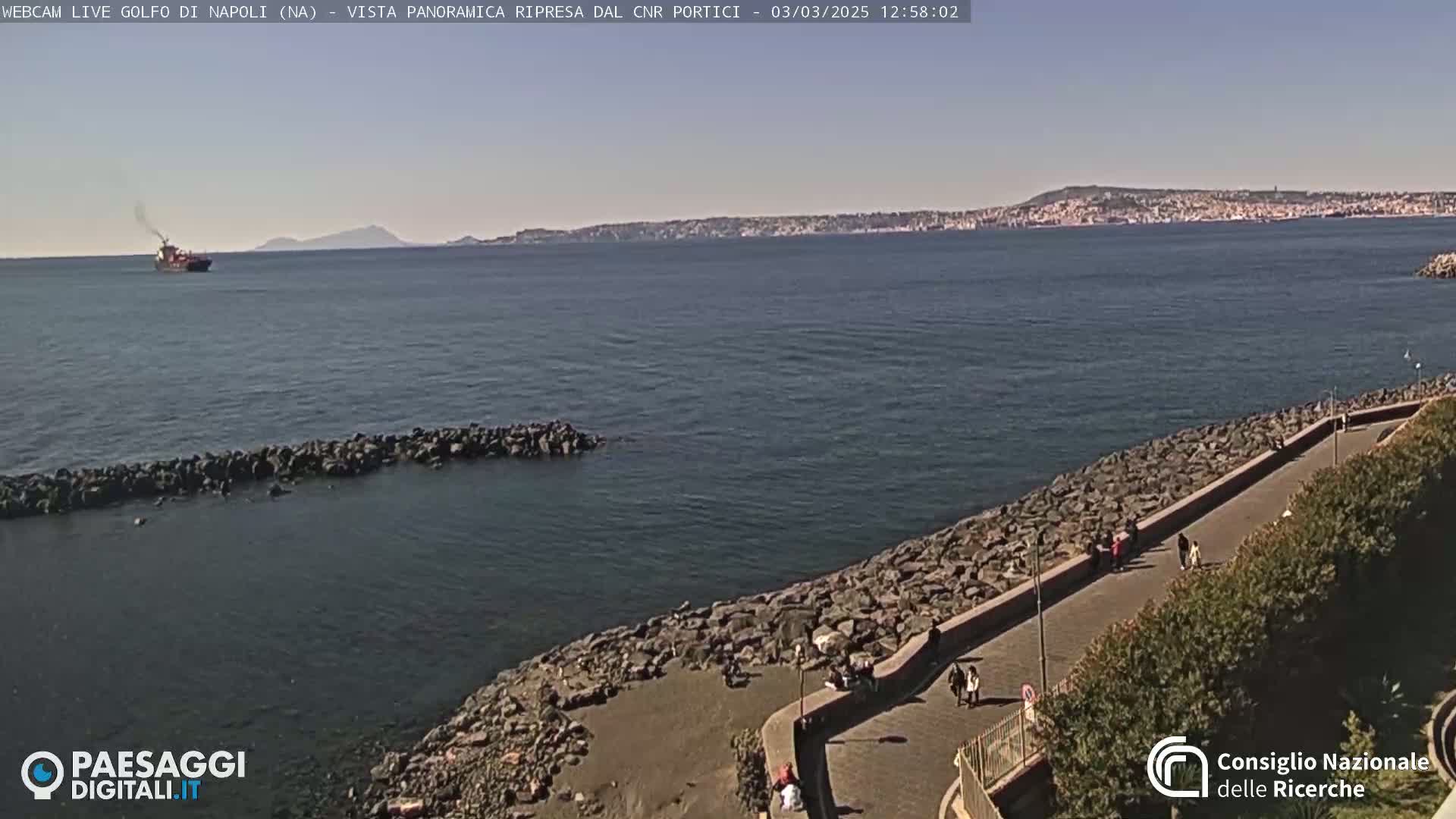A calm, sunny day shows a coastal path alongside a rocky shoreline, overlooking a body of water with a distant city and a small island visible.
